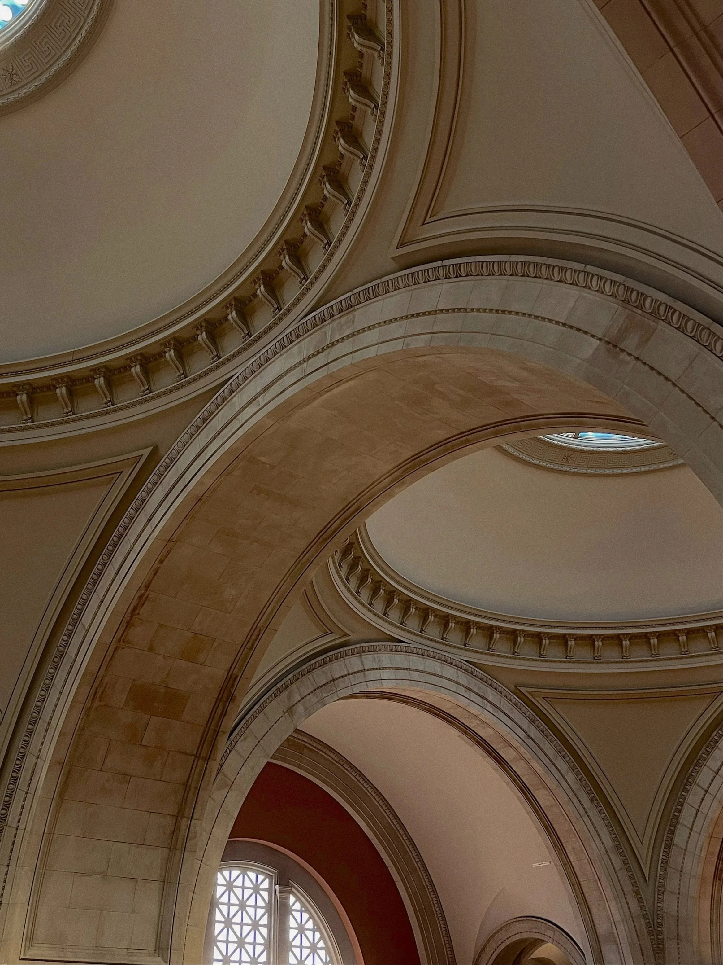 Interior view of an elegant, classical building with ornate arches, decorative moldings, and a window with a geometric lattice pattern.