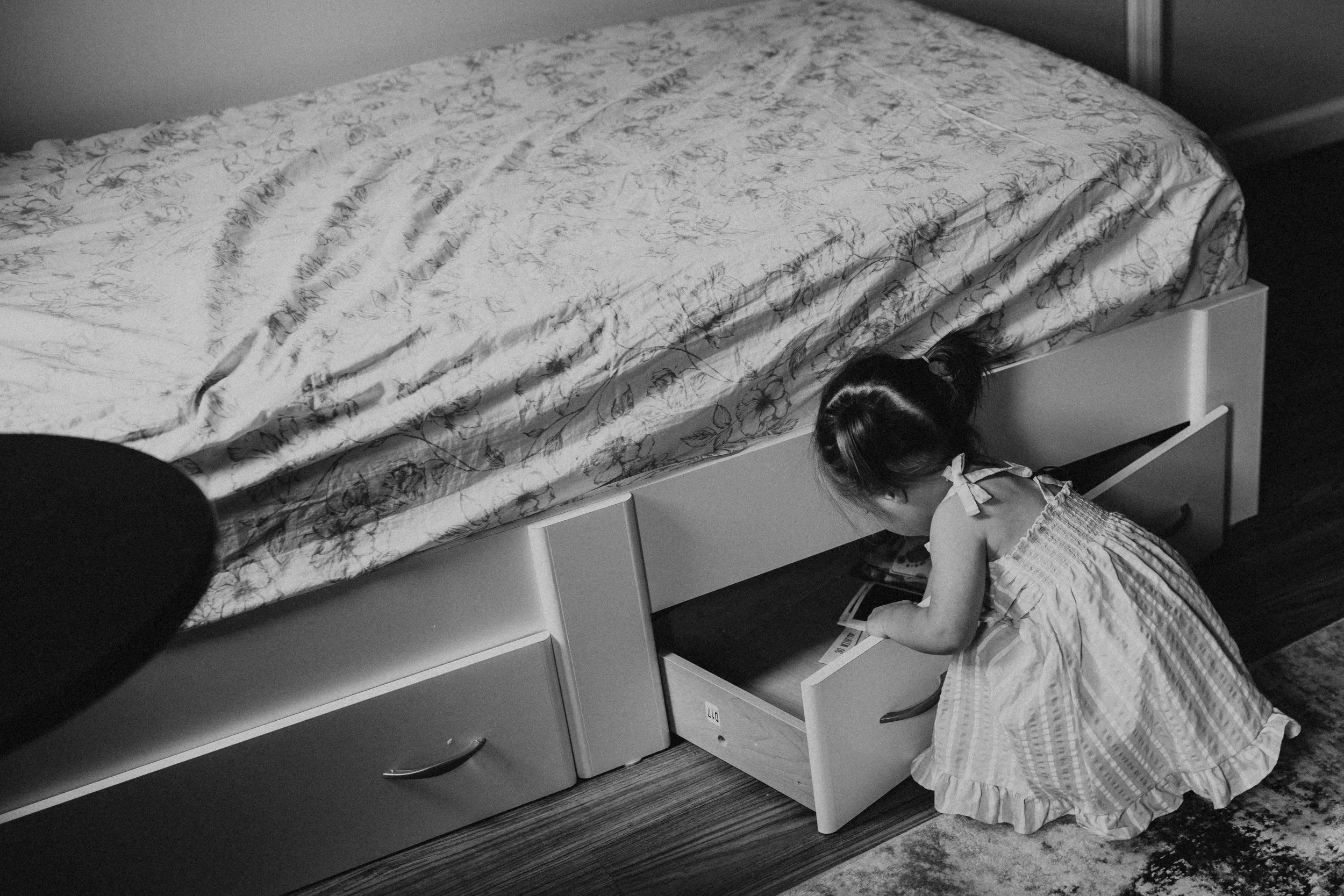 Girl in a dress reading a book while kneeling on the floor, partially hidden behind a piece of furniture with a floral bedspread on top.