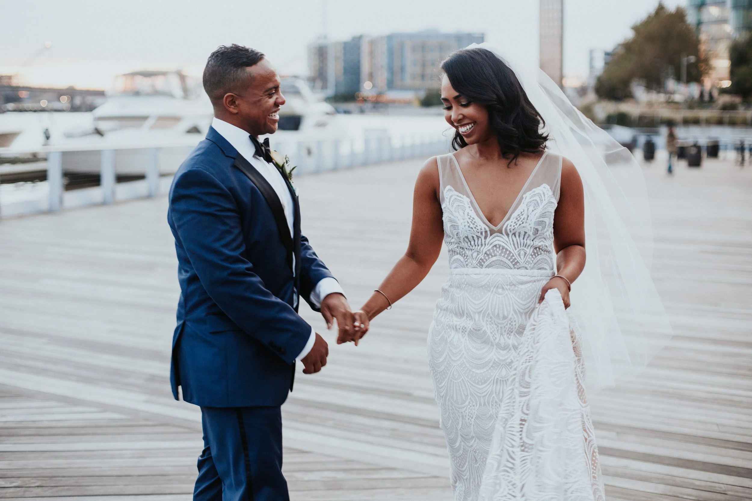 A bride and groom holding hands and smiling at each other outdoors near a marina, with boats and city buildings in the background.