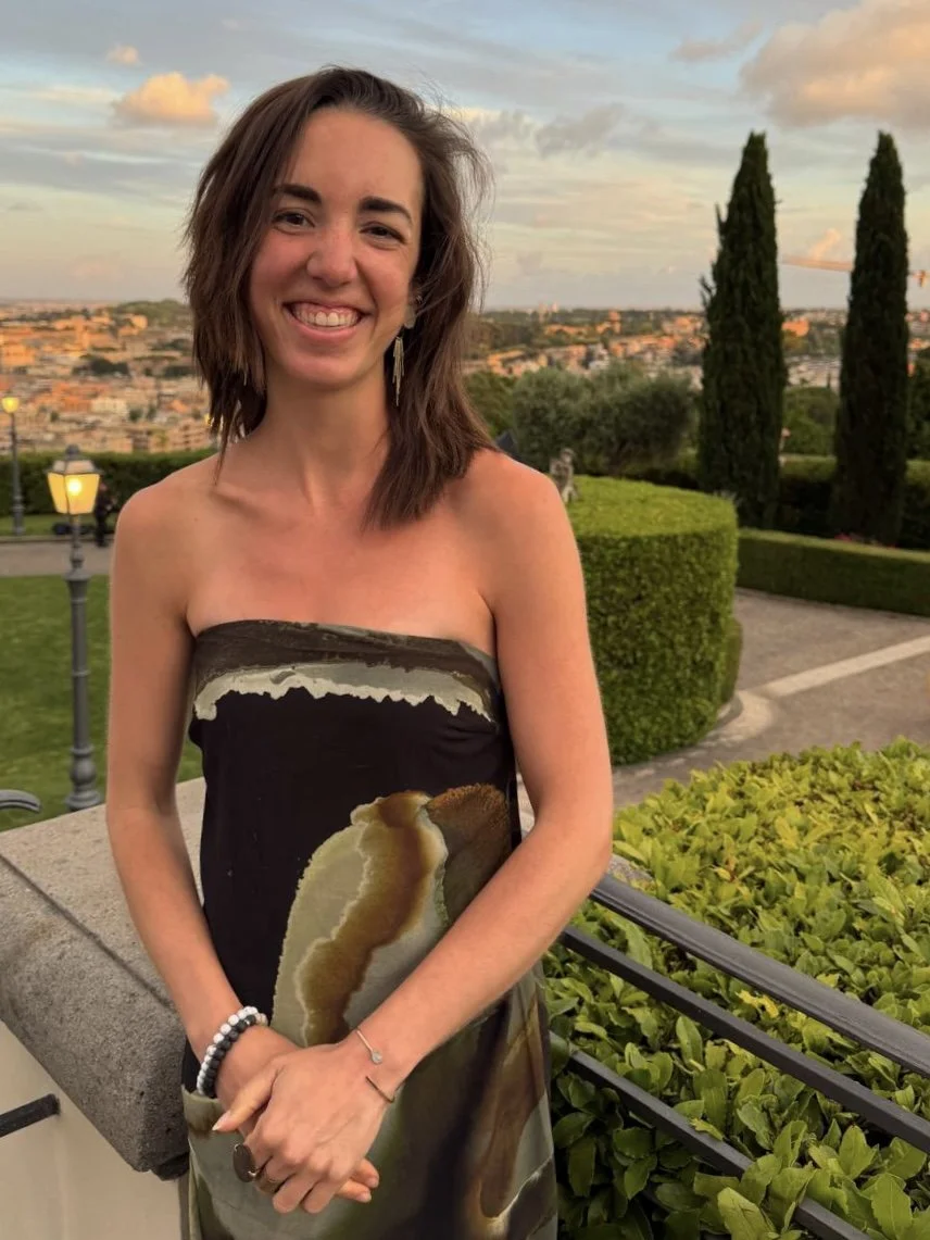 A woman standing outdoors at sunset, wearing a strapless dress with a scenic design, smiling with her arms crossed, surrounded by greenery and a cityscape in the background.