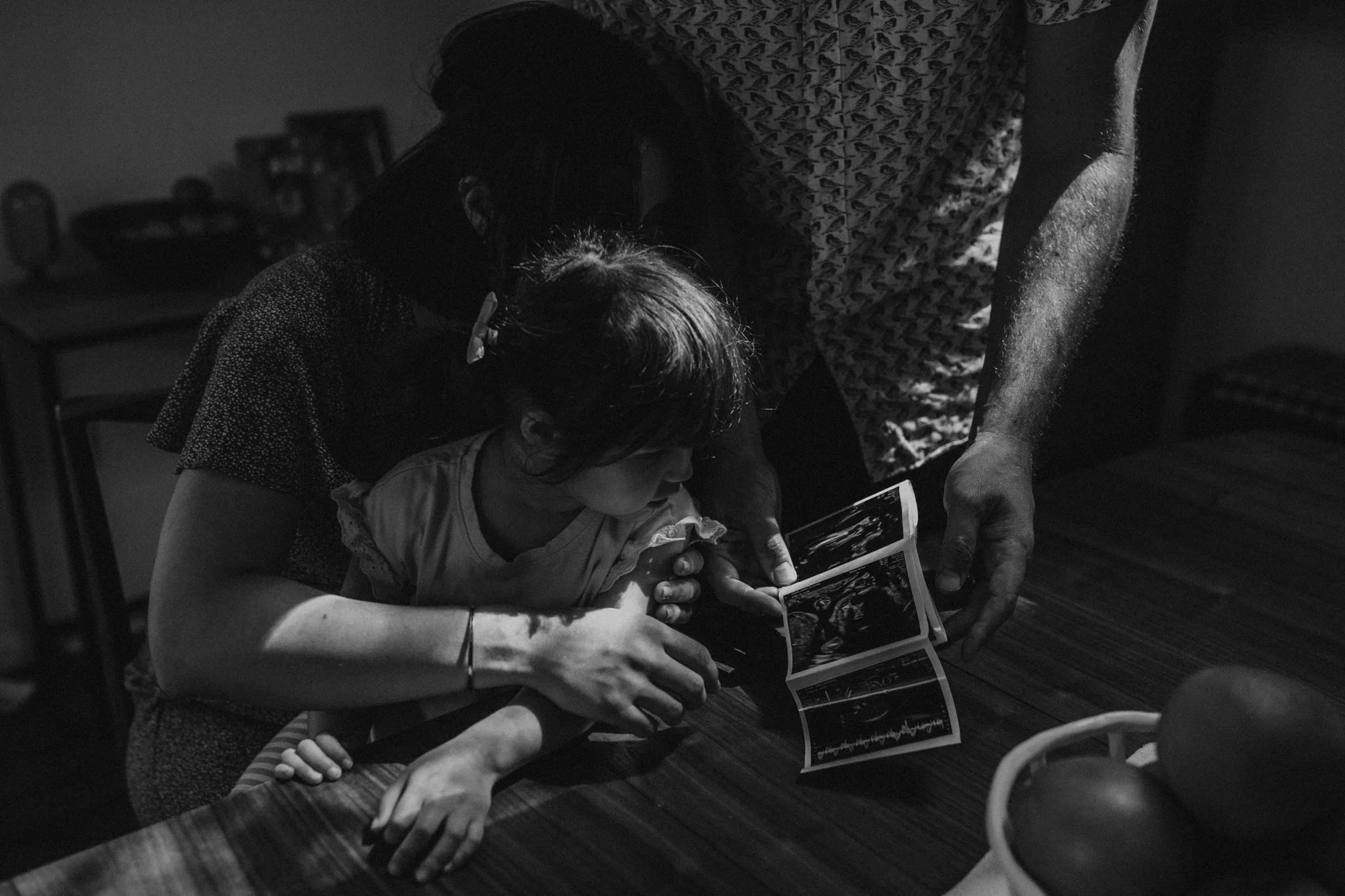 A woman and a young girl are looking at ultrasound images on a tablet. The woman is wearing glasses and a patterned shirt, and the girl is sitting on her lap with a wristband. They are in a dimly lit room, possibly a medical or examination room, with