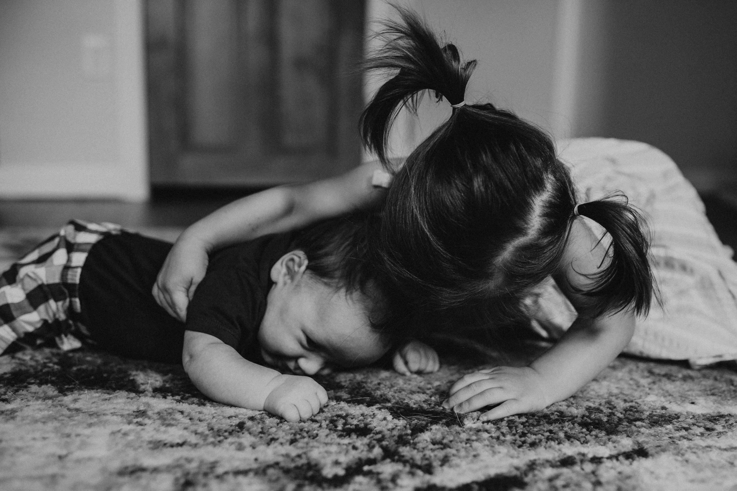 Two children, a girl with pigtails and a boy, are playing on a carpeted floor, with the girl lying on top of the boy, both laughing and enjoying their play.