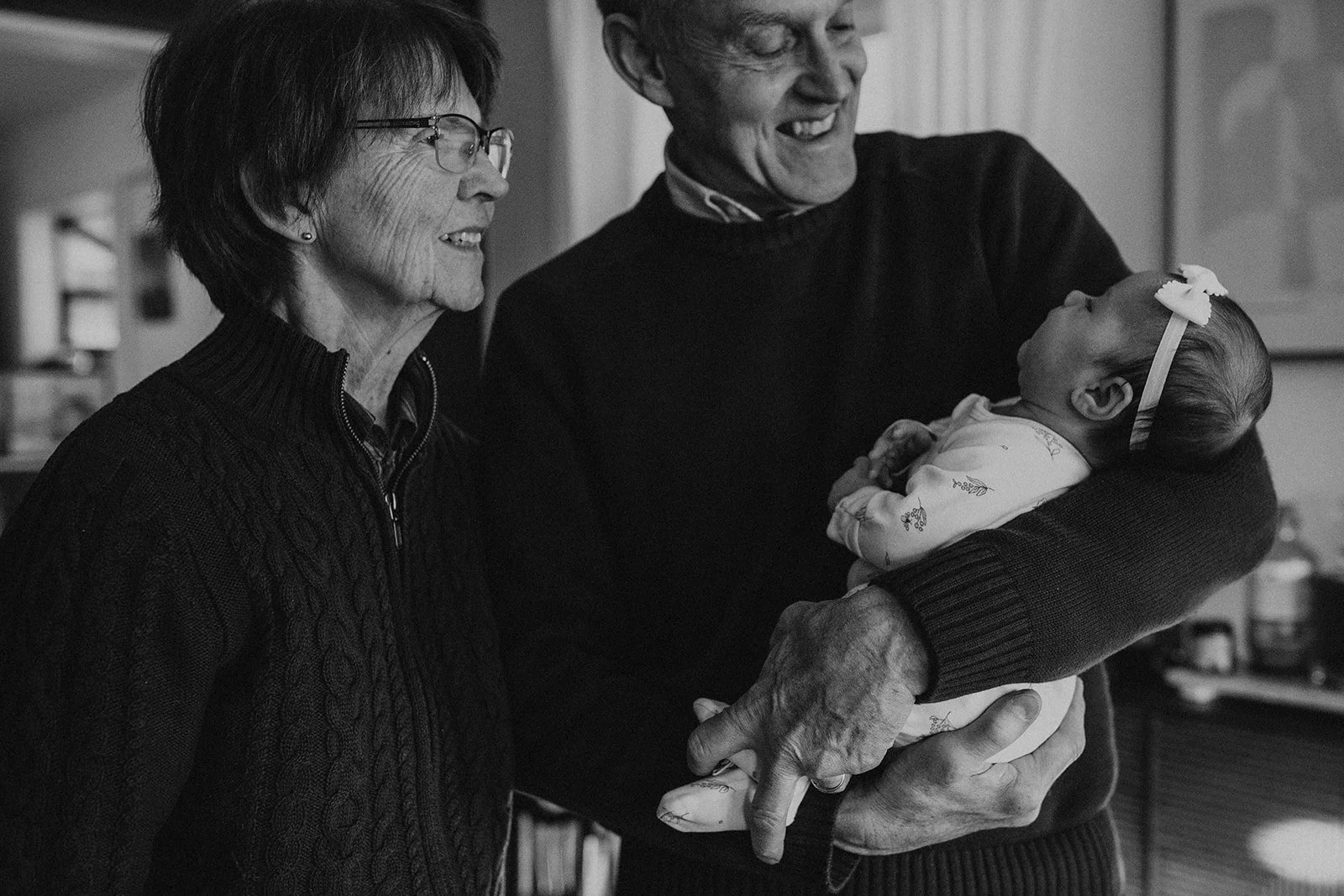 An elderly woman and man, both smiling, are looking at a baby girl being held in the man's arms. The woman has short hair, glasses, and is wearing a dark sweater, while the man has gray hair and is wearing a dark sweater. The baby girl is dressed in 
