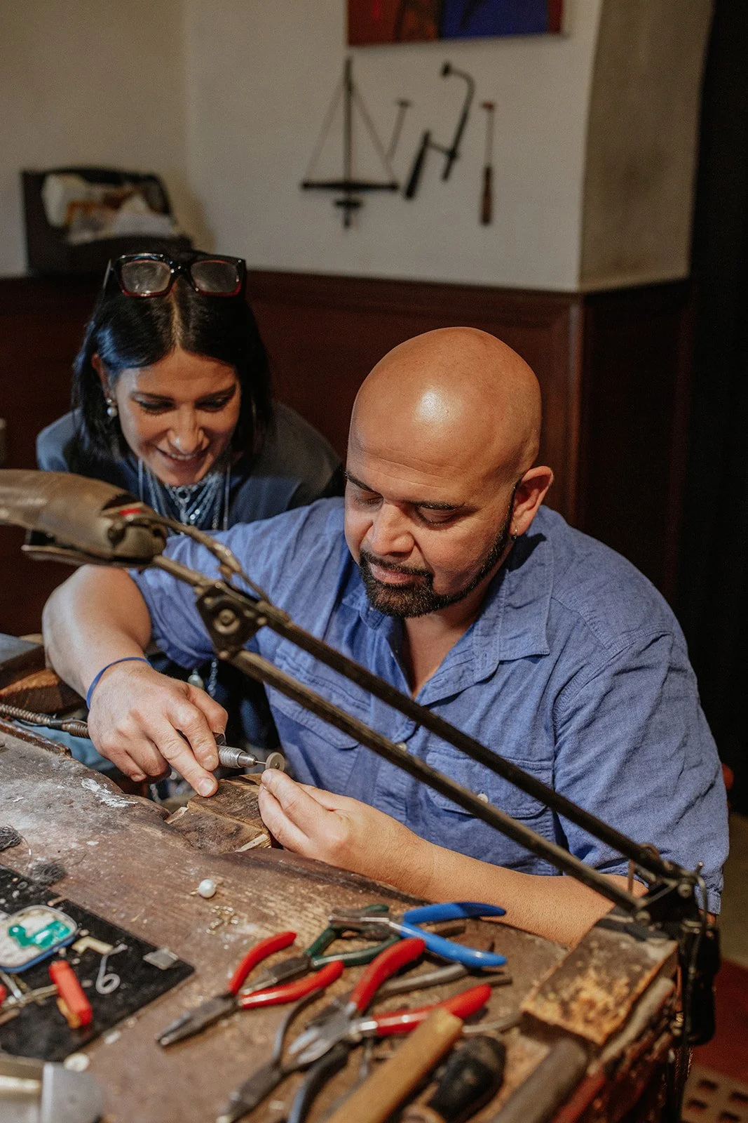 Two people, a man and a woman, working on a electronics project at a cluttered workbench, with tools and electronic components. The woman is smiling while watching the man, who is concentrating on using a tool.