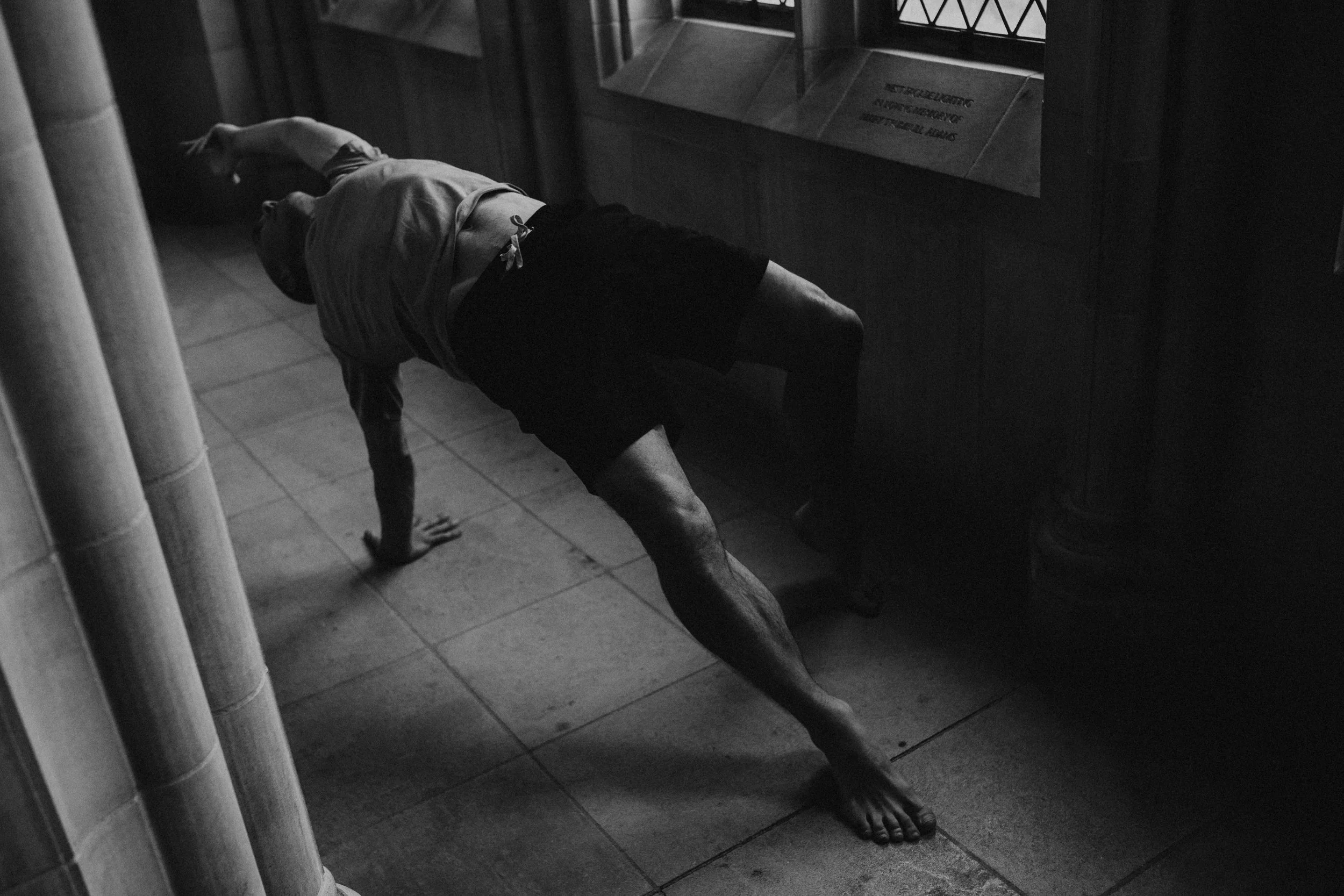 A man stretches in a yoga pose with their hand on the ground, wearing a t-shirt and shorts, in a dimly lit ancient church hallway.