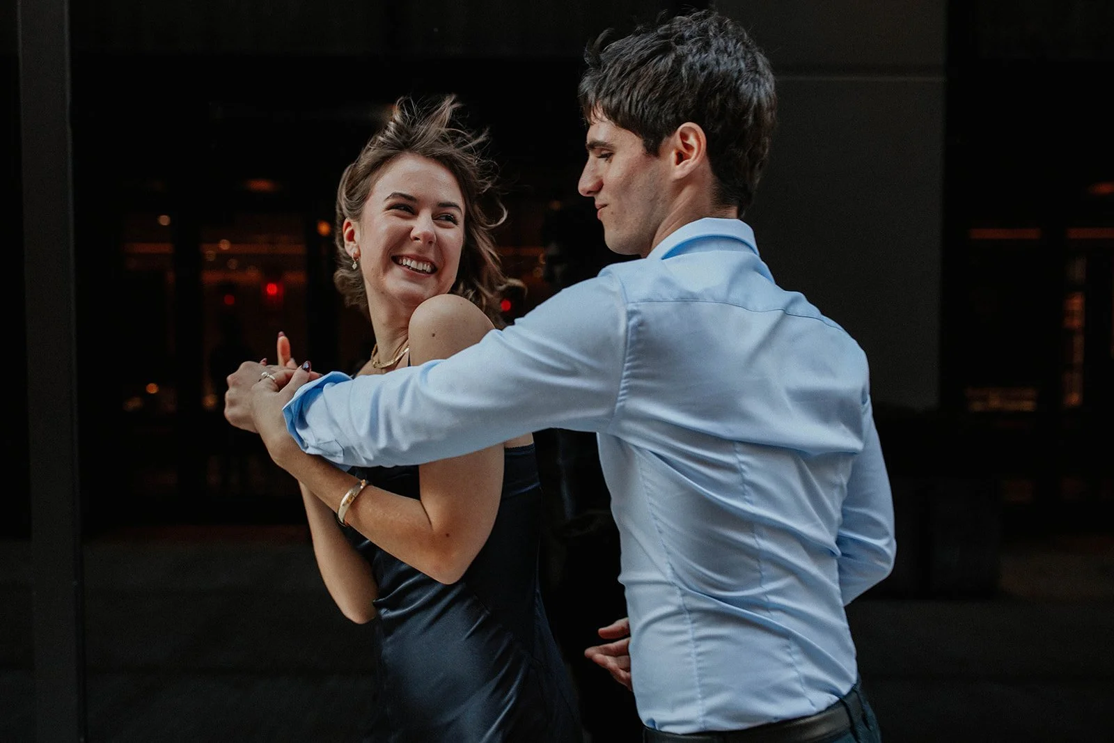 A young woman with short brown hair smiling and holding hands with a young man with dark hair, wearing a blue shirt, while dancing together indoors at night.