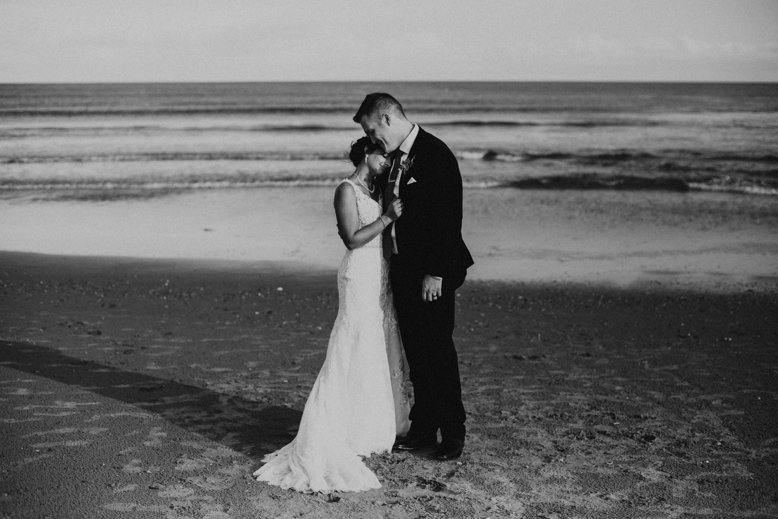 Black and white photo of a couple in wedding attire embracing on a sandy beach near the ocean, with waves in the background.
