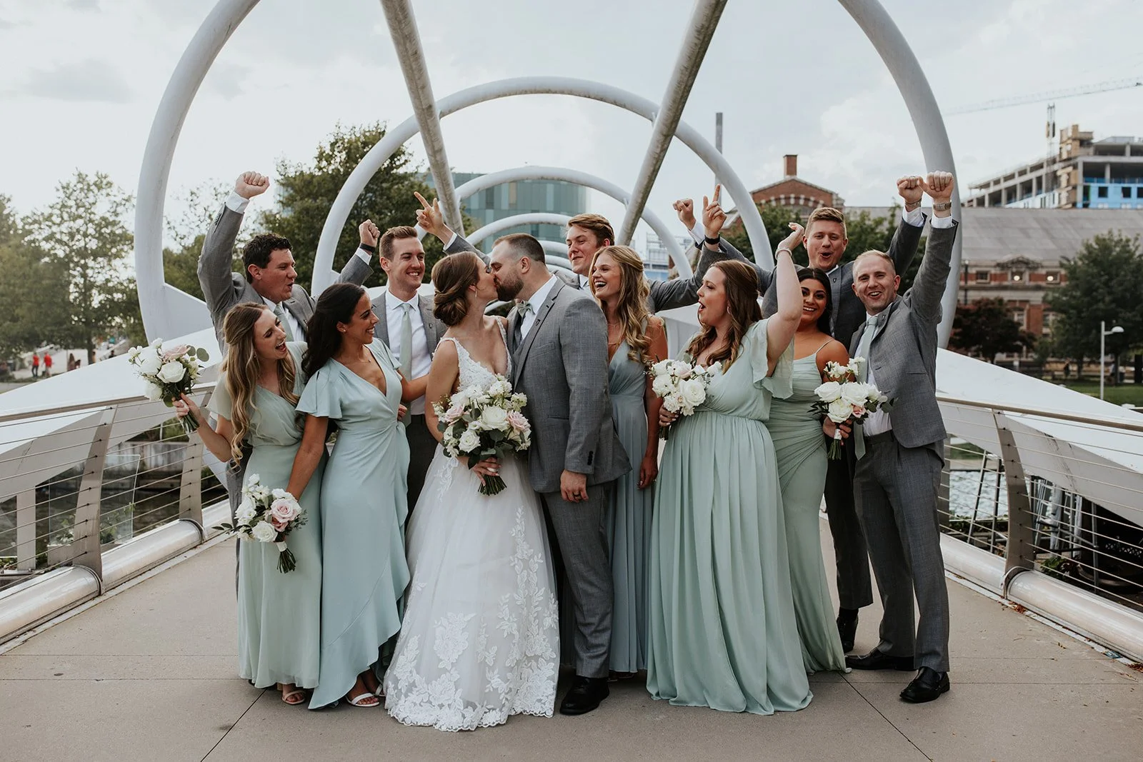 A wedding party with the bride and groom kissing, surrounded by bridesmaids and groomsmen celebrating on a modern bridge outdoors.