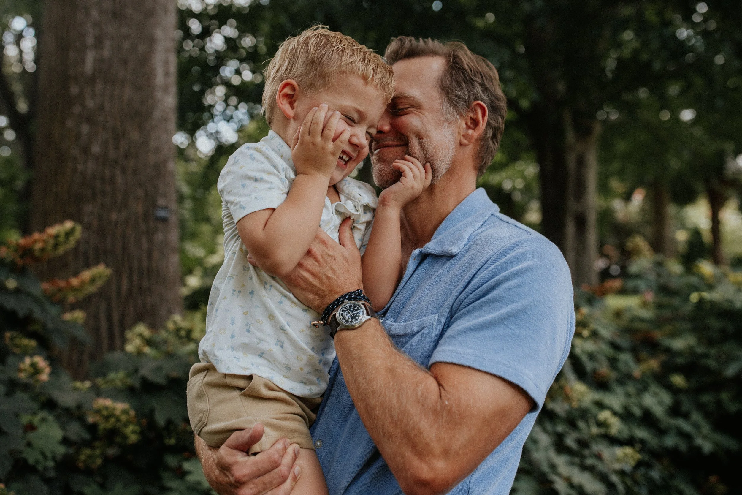 A man holding a young boy, both smiling and laughing, in a forested park.