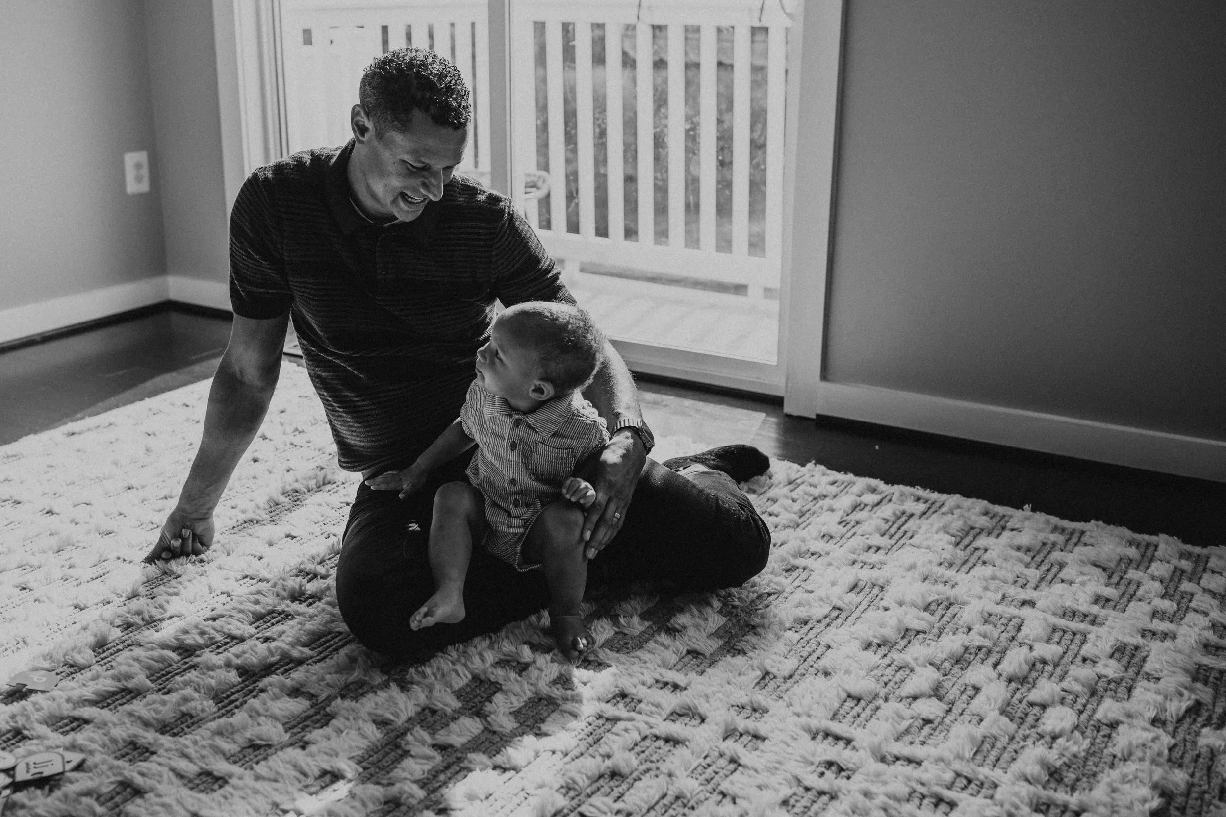 A black and white photo of a man and a young boy sitting on a carpet, smiling and looking at each other. The man is on the floor, leaning on one arm, with the boy sitting on his lap. The room has a large window with a wooden balcony railing outside. 
