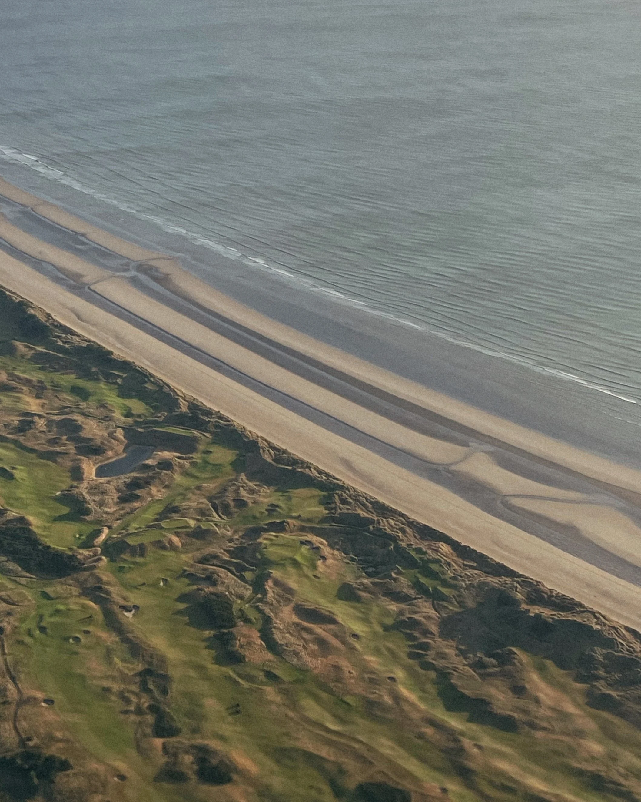 Aerial view of a coastline with a beach, ocean, and inland greenery.