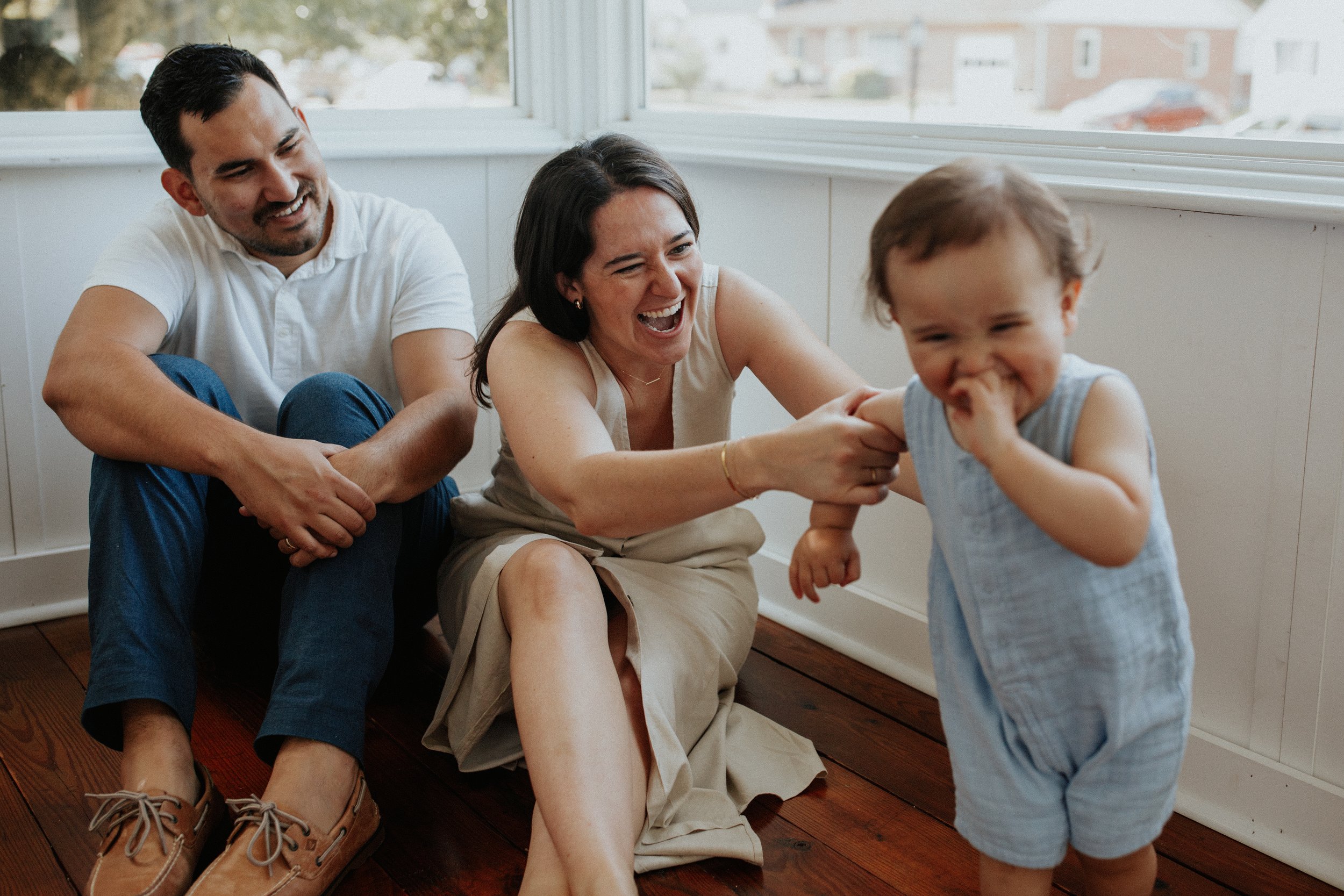 A happy family playing together indoors near a large window. A man and woman are sitting on the floor, laughing, while a young boy in a striped outfit stands and smiles.
