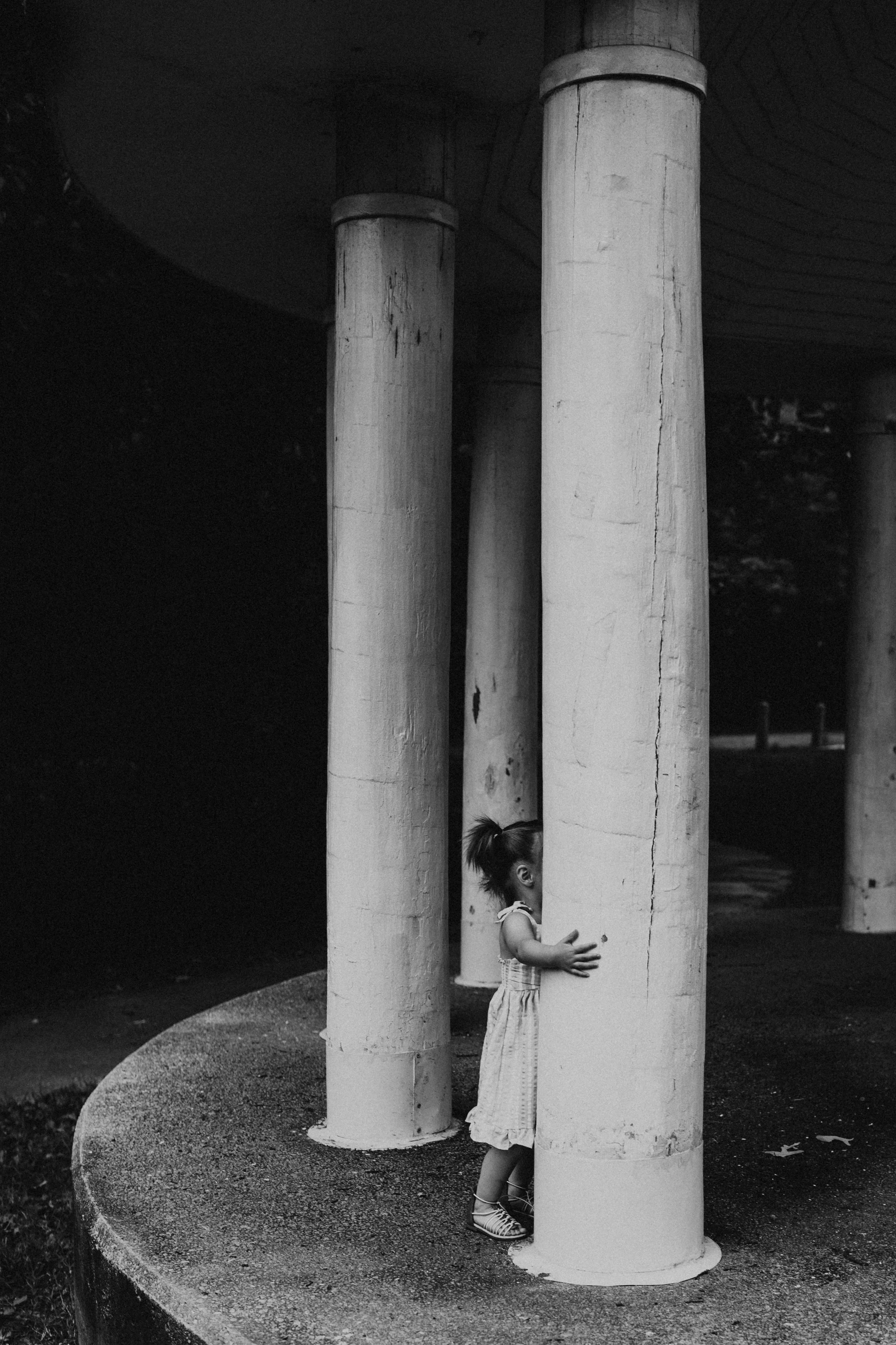 A young girl with pigtails wearing a dress and striped shoes stands behind a large concrete pillar, peering around it with her head and hands visible. There are several similar concrete pillars in the background, and the scene appears to be outdoors 