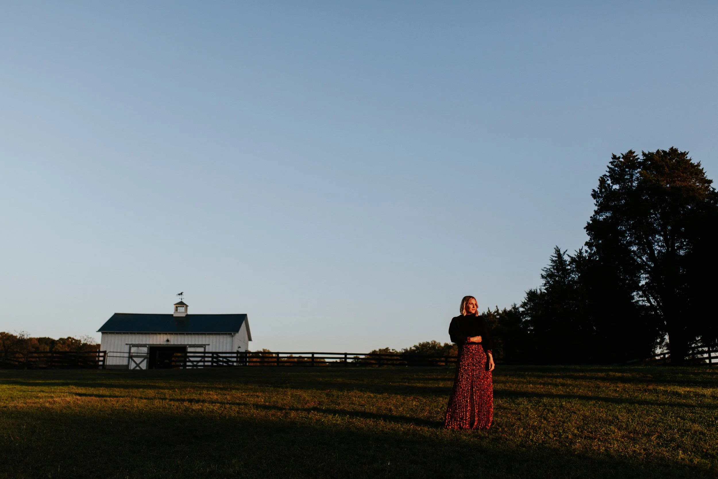 A woman in a long, patterned dress stands outdoors during sunset or twilight, near a large tree and a white building with a black roof and a weather vane on top. The sky is clear and faintly illuminated.
