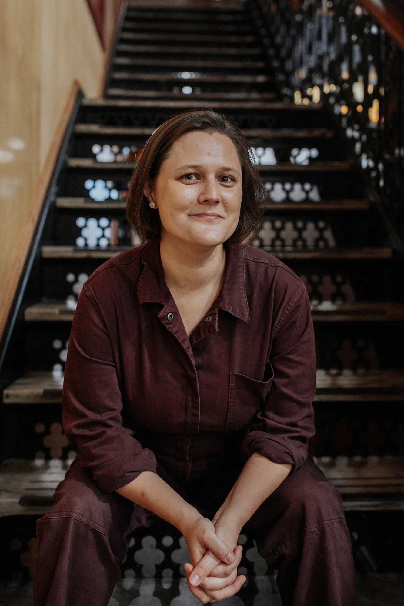 A woman with short brown hair and wearing a burgundy shirt sitting on stairs in front of a decorative railing.
