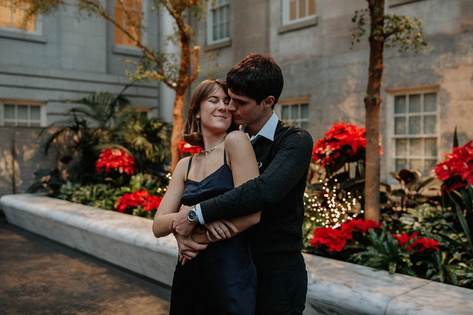 A couple hugging at an outdoor evening event decorated with red poinsettias and string lights, with a building and trees in the background.
