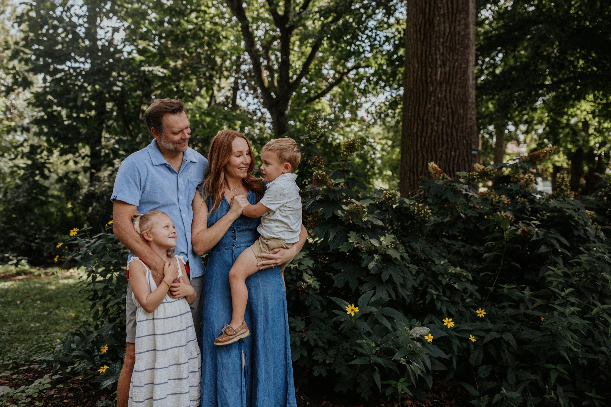 A family of four, including a man, woman, young girl, and young boy, standing outdoors in a lush green area with trees and yellow flowers. The woman is holding the boy, and they are all smiling and enjoying the moment.