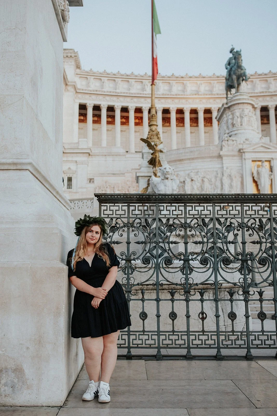 A young woman in a black dress and white sneakers standing outdoors near a decorative metal gate, with a historical monument and Italian flag in the background.
