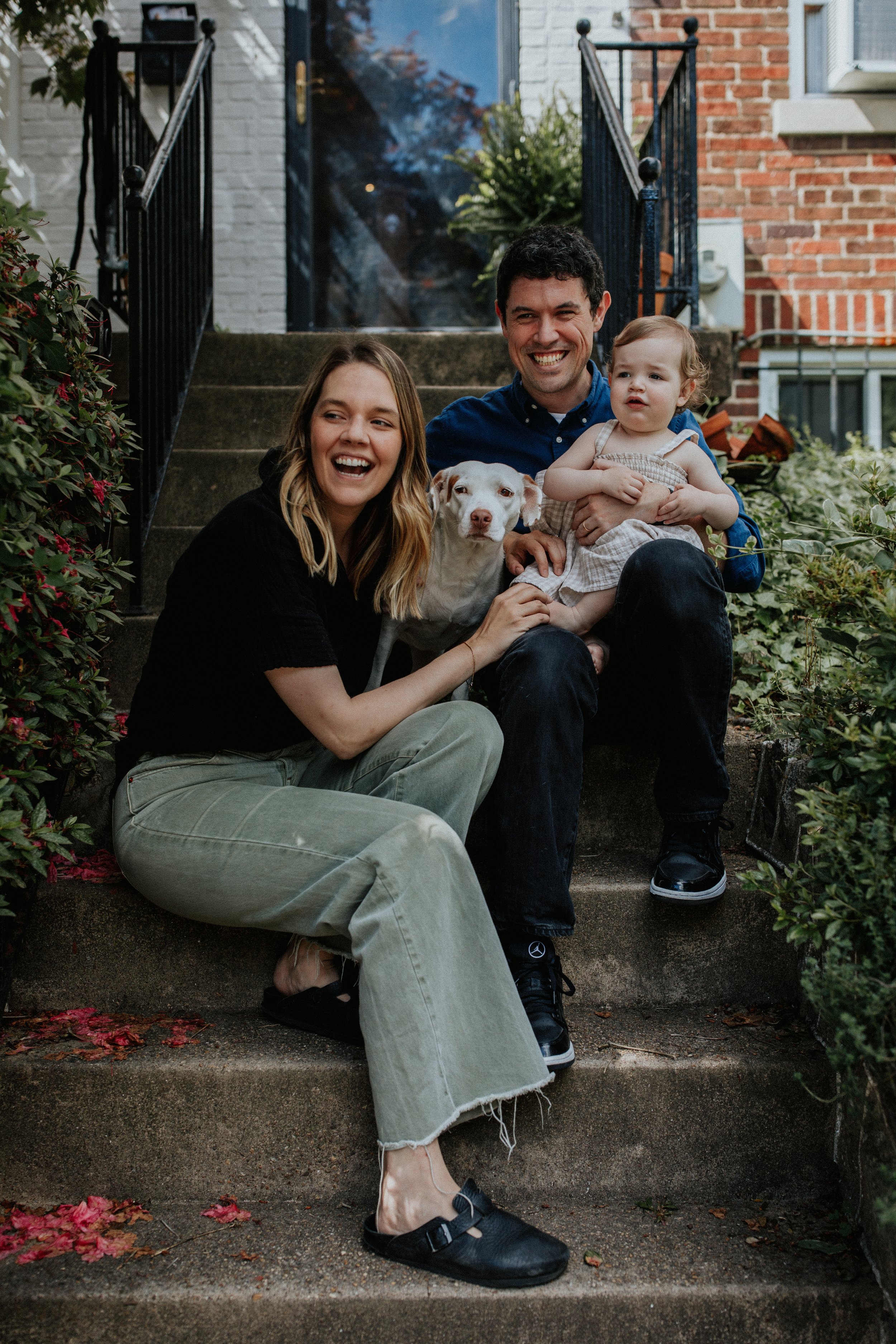 A family sitting on front steps of a house, smiling and holding a dog, with a large dog's face on a glass door in the background. The family includes woman with blonde hair, man with dark hair, and a young child with curly blonde hair, surrounded by 