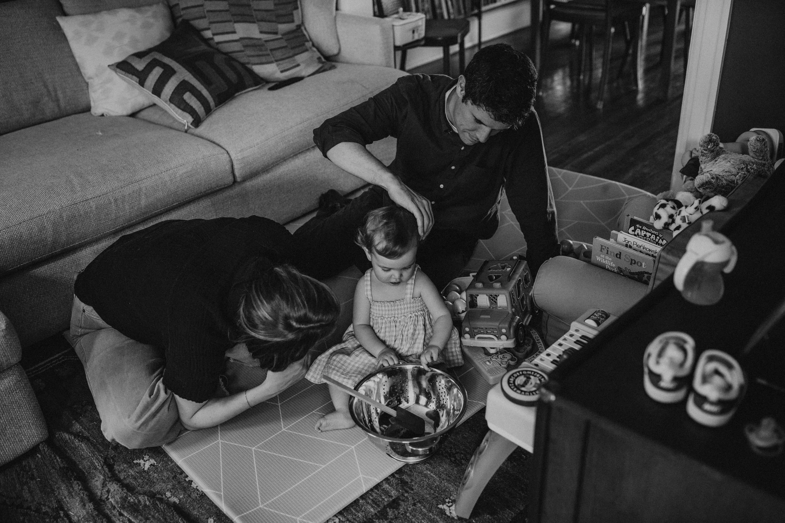 A man and woman are playing with children and toys on the floor in a living room. The children are sitting among toys, and the man is touching one of the children. There is a sofa with pillows, a bookshelf, and a stool in the background.