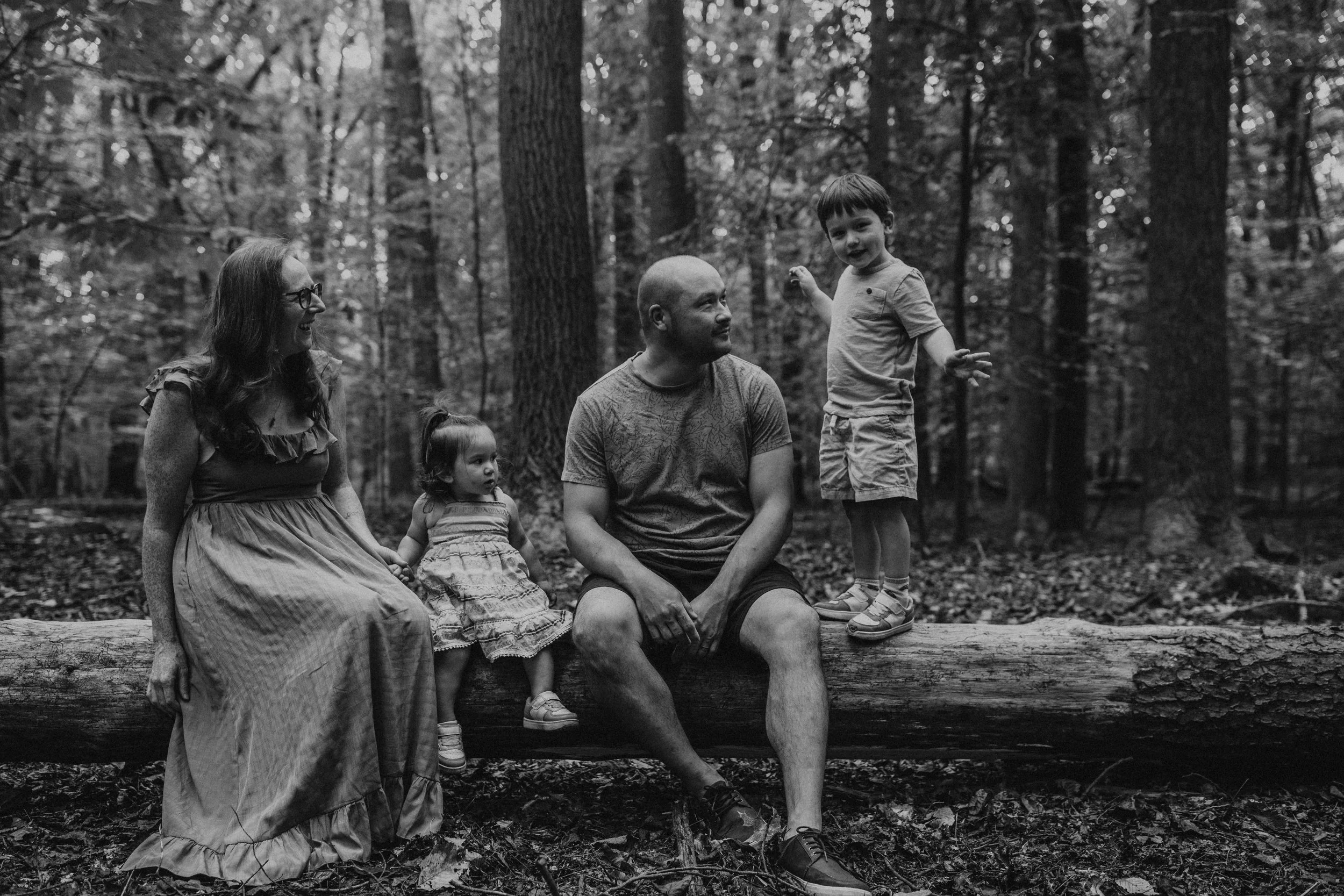 A black-and-white photo of a family in a forest, sitting and standing on a fallen log. There are three children and two adults. One child is standing and talking, while the others sit and listen.