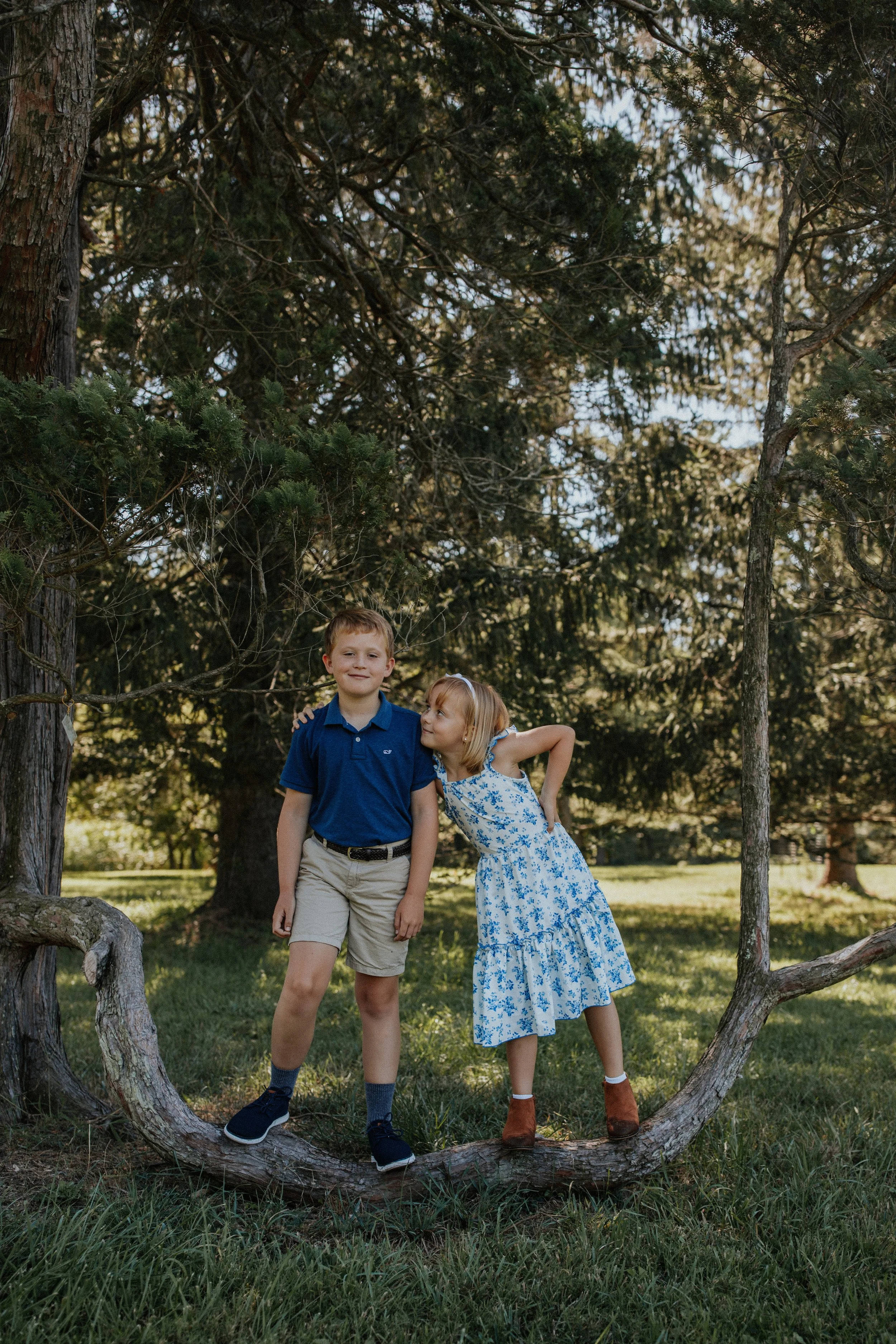 Two children, a boy and a girl, standing on a tree branch outdoors in a park with trees in the background.