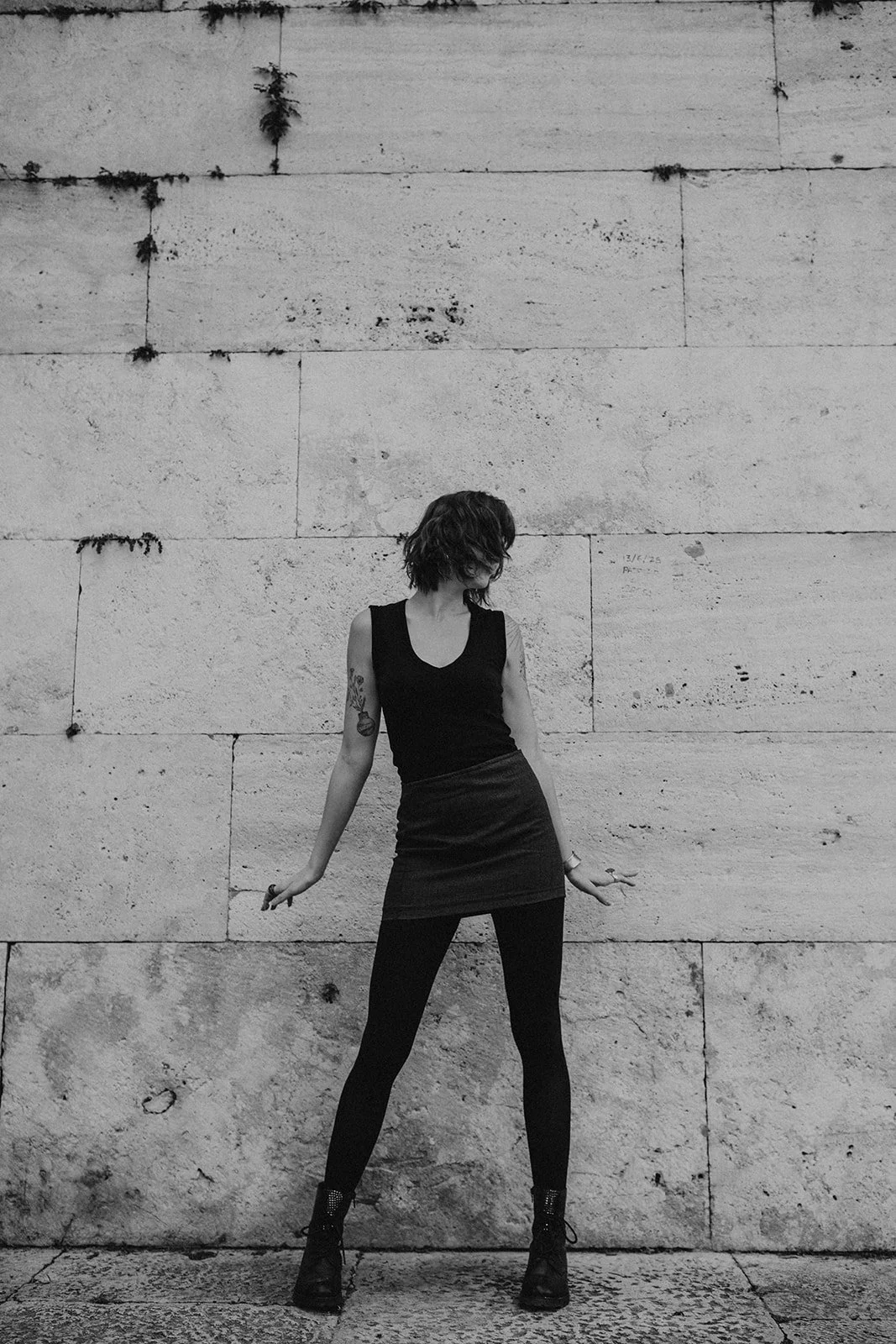 A woman with short curly hair and tattoos on her right arm poses against a large, textured stone wall, wearing a black sleeveless top, a dark skirt, black tights, and black combat boots.