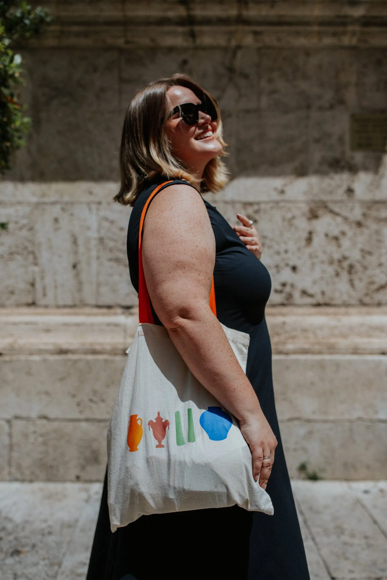 A woman wearing sunglasses and a black top, smiling and standing outdoors against a stone wall in Rome, Italy. She is holding a tote bag with colorful images of various objects on it.