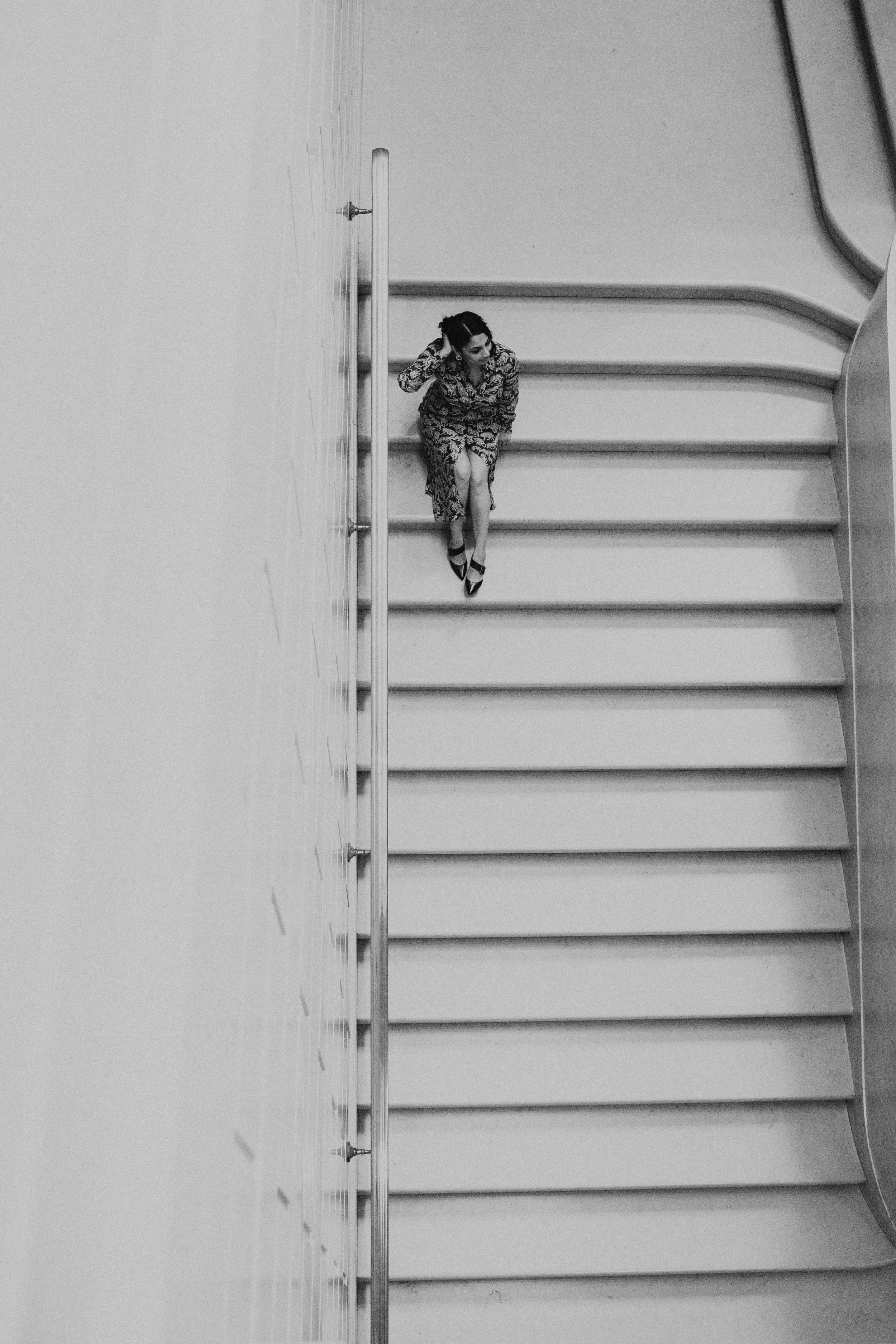Black and white photo of a woman sitting on a staircase, wearing a patterned dress and high heels, with her hand running through her hair.