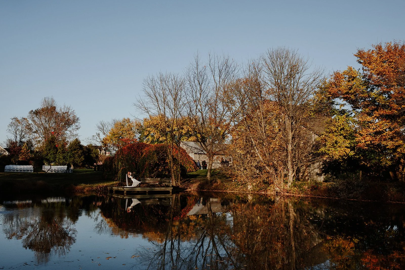 Autumn scene at a park with a pond, trees with fall foliage, a woman in a white dress sitting on a dock by the water, and houses in the background.