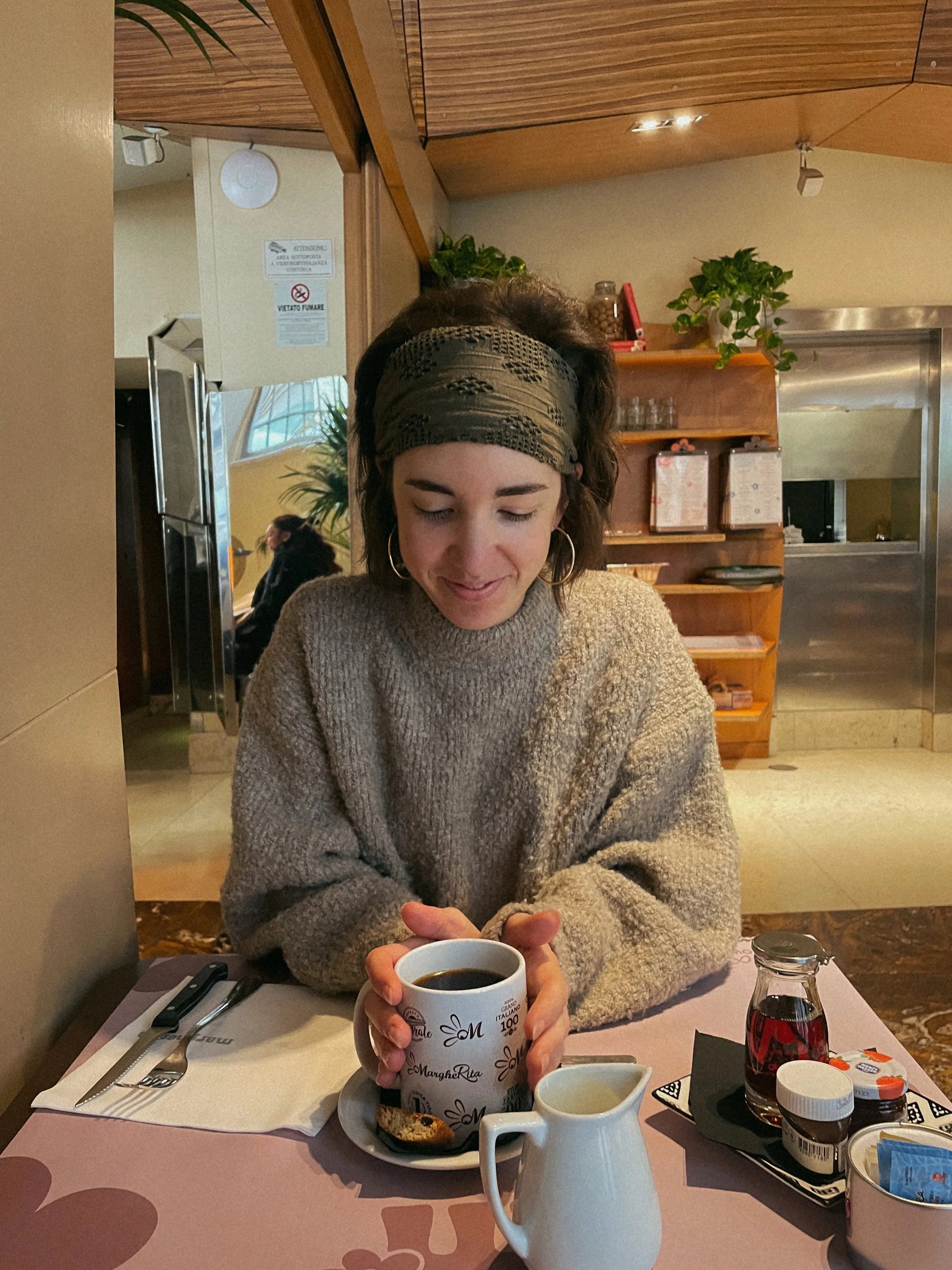 A woman sitting at a restaurant table holding a cup of coffee. The table has breakfast items, a glass of water, and condiments. The background has warm lighting, a wooden shelf with plants, and another woman seated behind her.