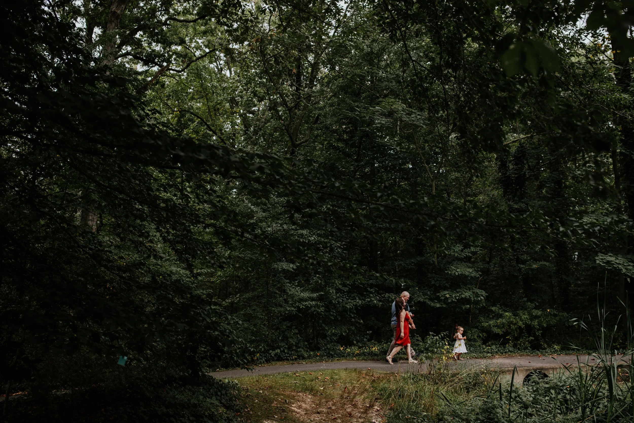 Three people walking along a path in a dense, green forest. The group includes an elderly man, a woman in a red dress, and a young girl wearing a white dress.