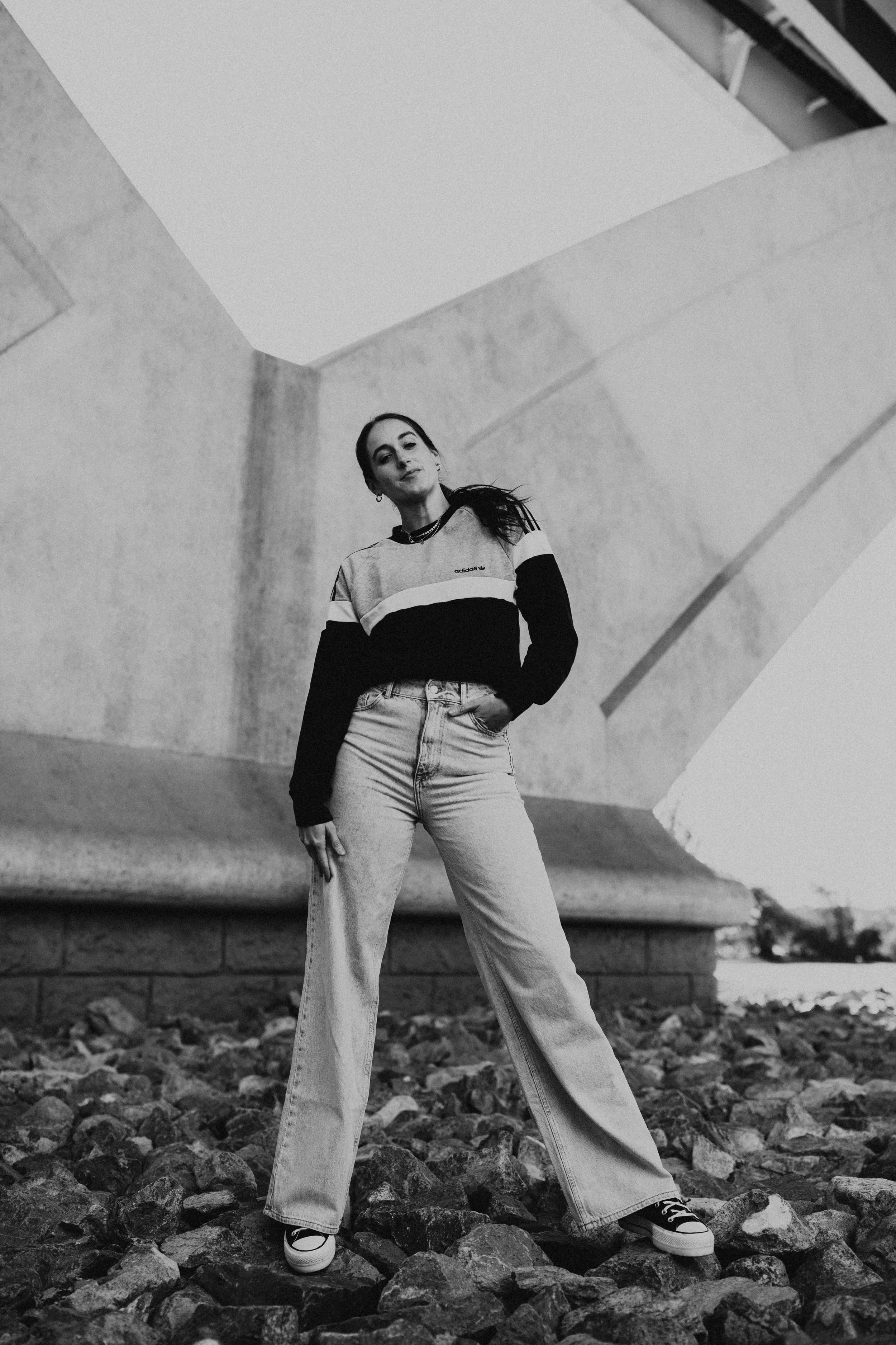 A woman with long dark hair, wearing a sweatshirt with stripes, jeans, and sneakers, standing on rocky ground under a bridge in black and white.