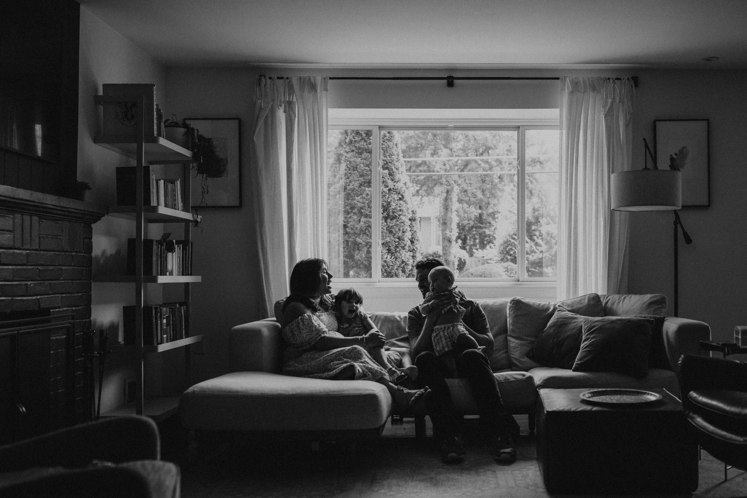 A living room with large window showing trees outside, a bookshelf, artwork, and a floor lamp. Four children are sitting on a sofa, with one holding a baby, interacting and smiling.