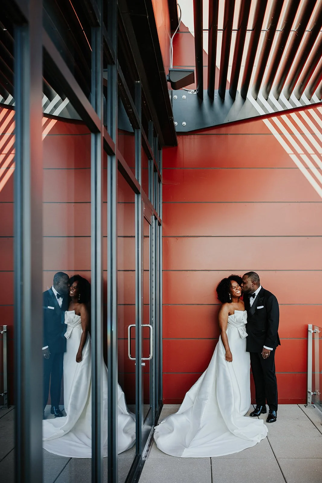 A couple in wedding attire sharing a kiss and laughing outside a modern building with red walls and glass reflections.