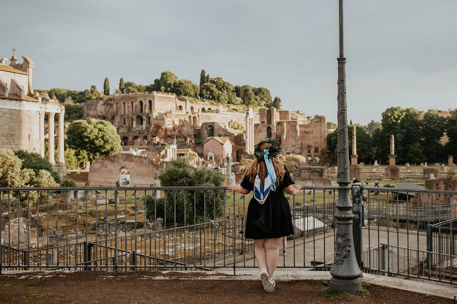 A woman with long hair wearing a black dress and a floral crown with blue and white ribbons stands in front of an ancient archaeological site, looking out over ruins and old structures.