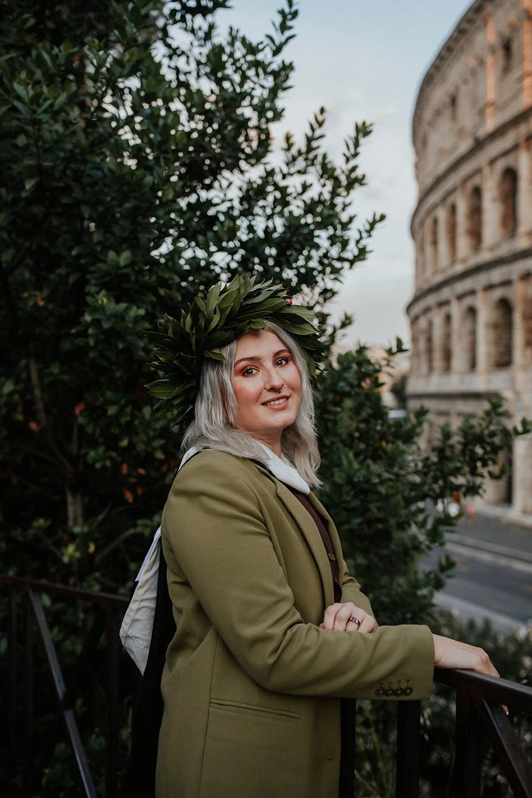 A young woman with silver hair, wearing a green laurel wreath, green jacket, and white shirt, standing outdoors near the Colosseum in Rome, Italy.
