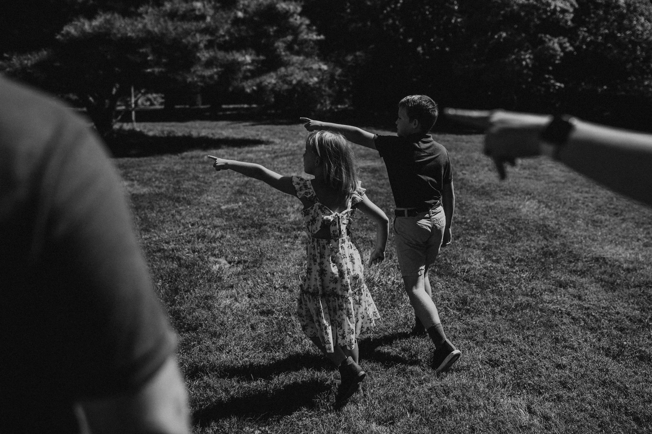 Two children, a girl and a boy, pointing into the distance while standing on grass in a park, with trees in the background, in a black and white photo.