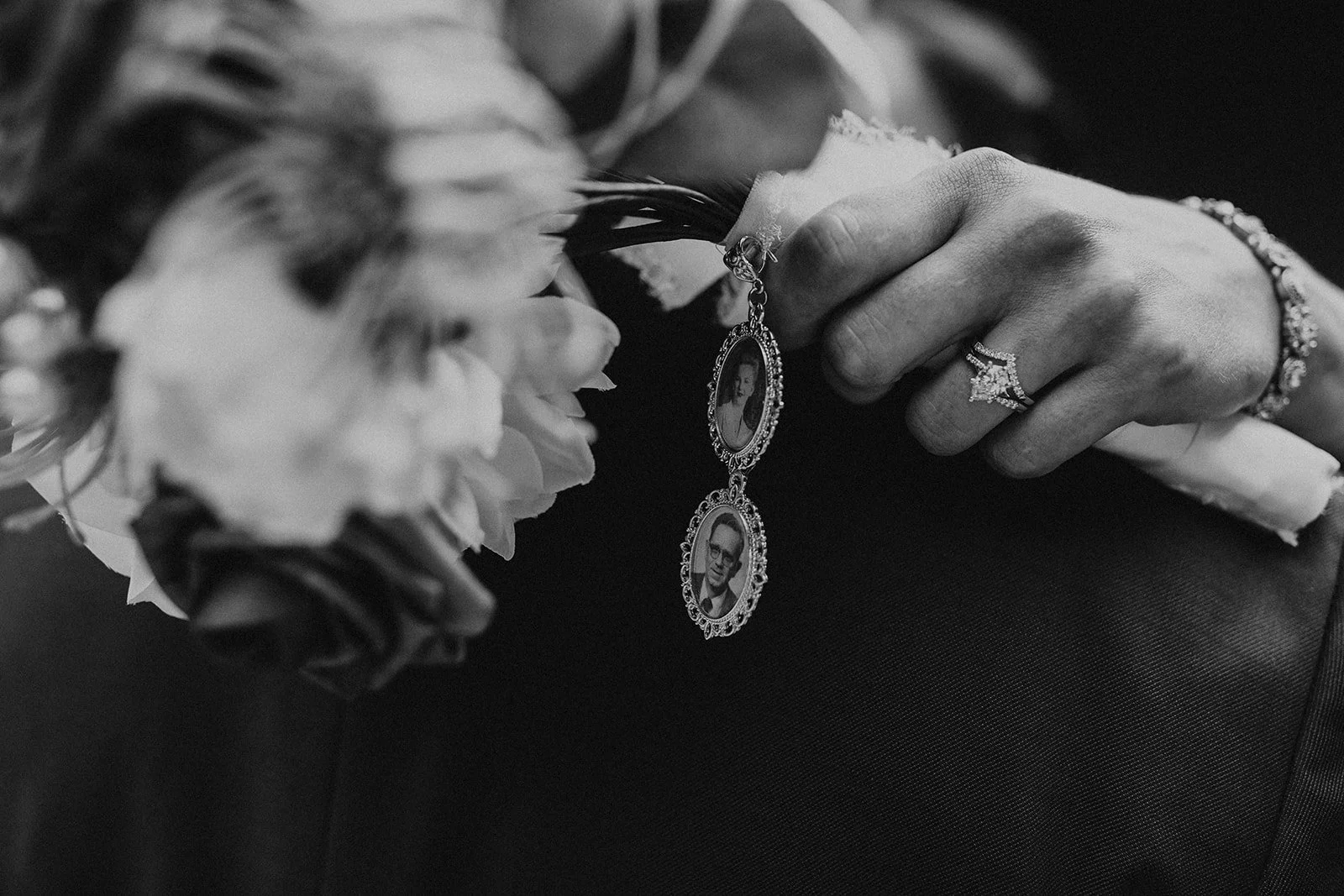 Close-up of a hand with a diamond ring and a bracelet, holding a pendant with two black-and-white photographs, resting on a bouquet of flowers.