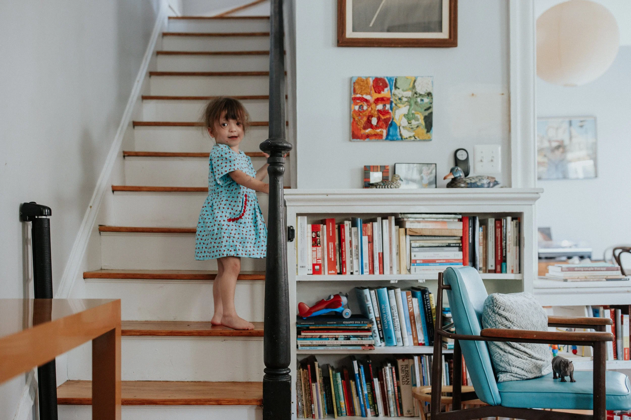 A young girl with brown hair and a blue dress with red polka dots stands barefoot on a staircase, holding a black stair railing, in a home filled with books and artwork.