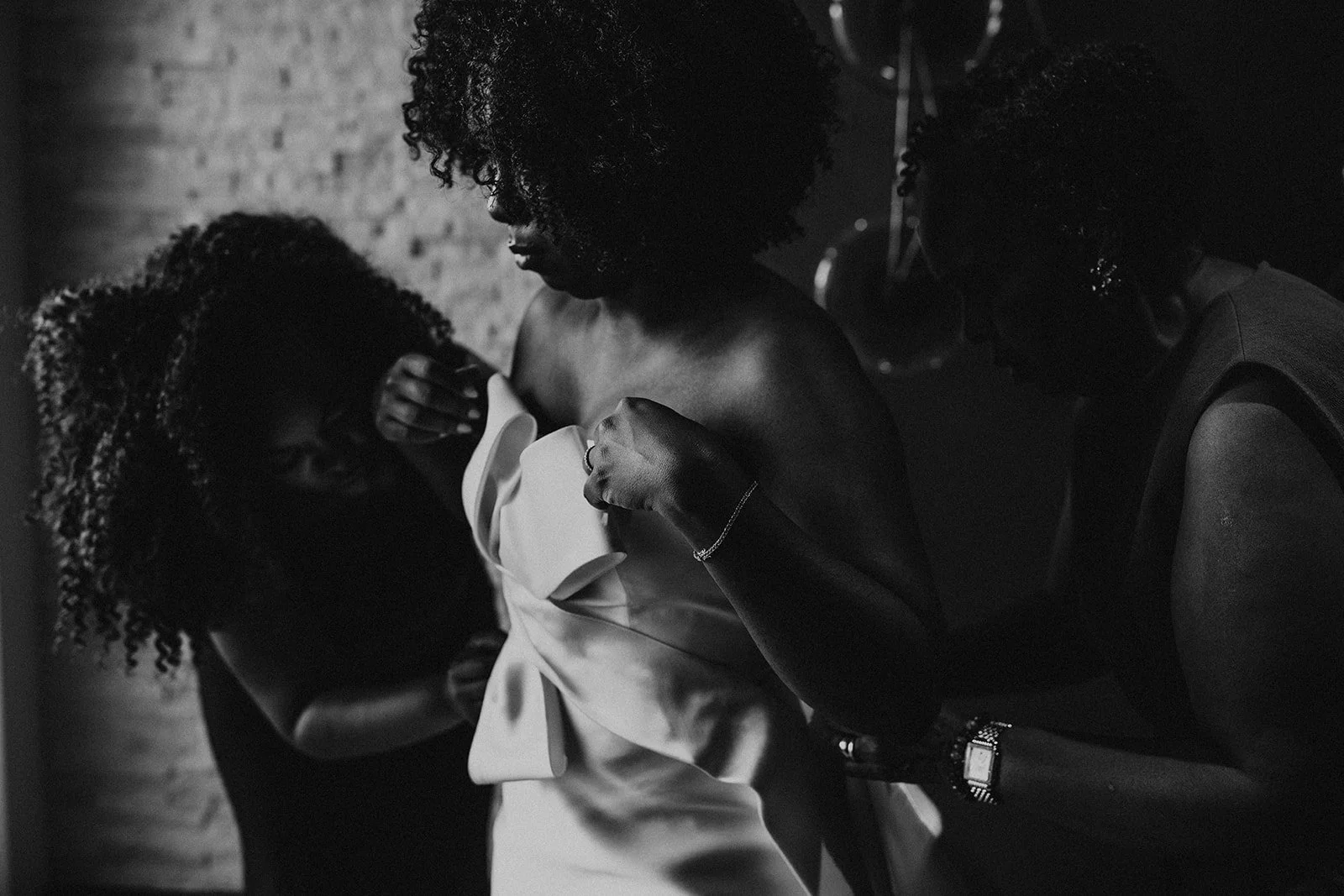 Three women in a dance studio, with two women helping a woman put on or adjust a wedding dress, in black and white photography.