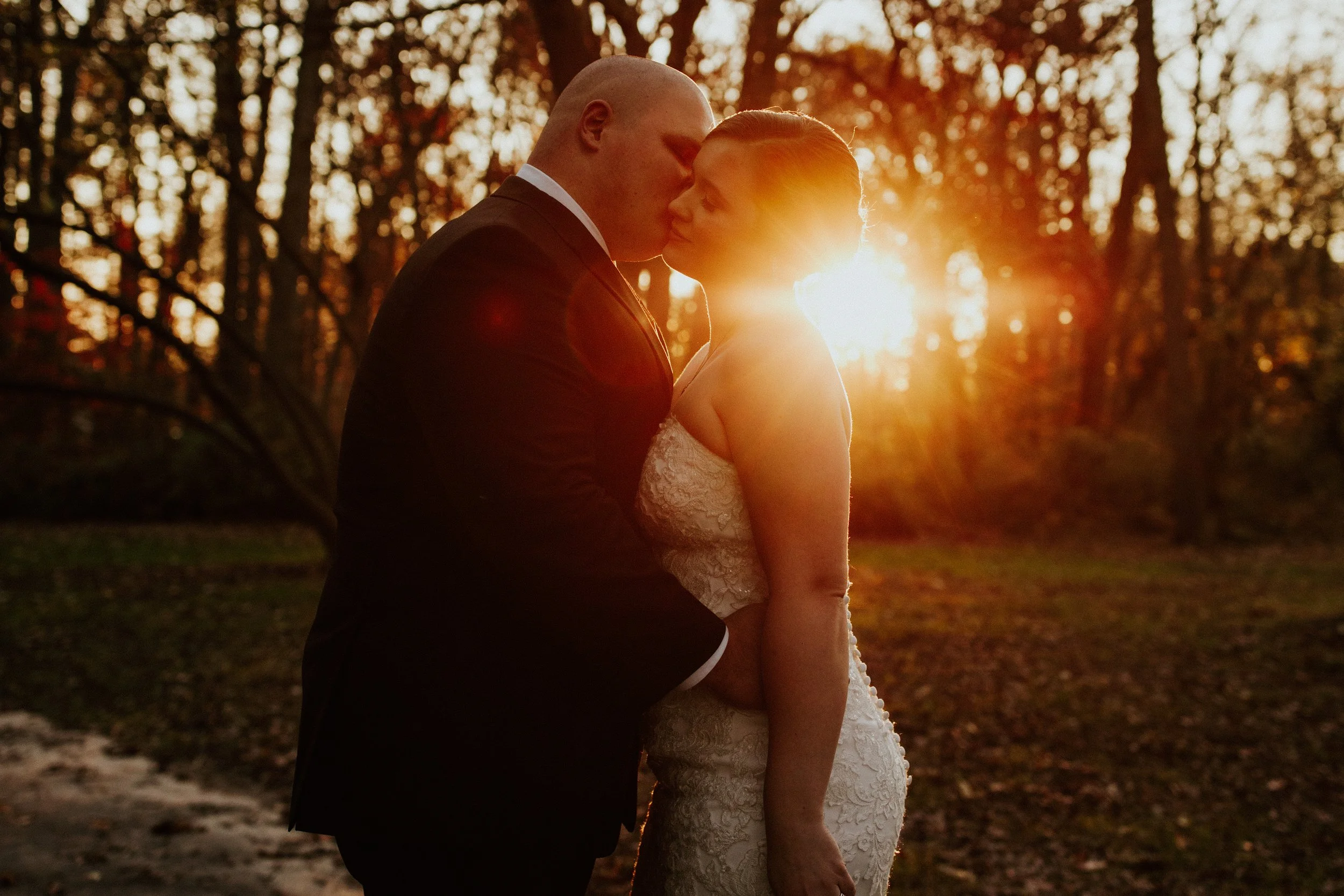 A couple dressed in wedding attire sharing a kiss outdoors during sunset in a wooded area.