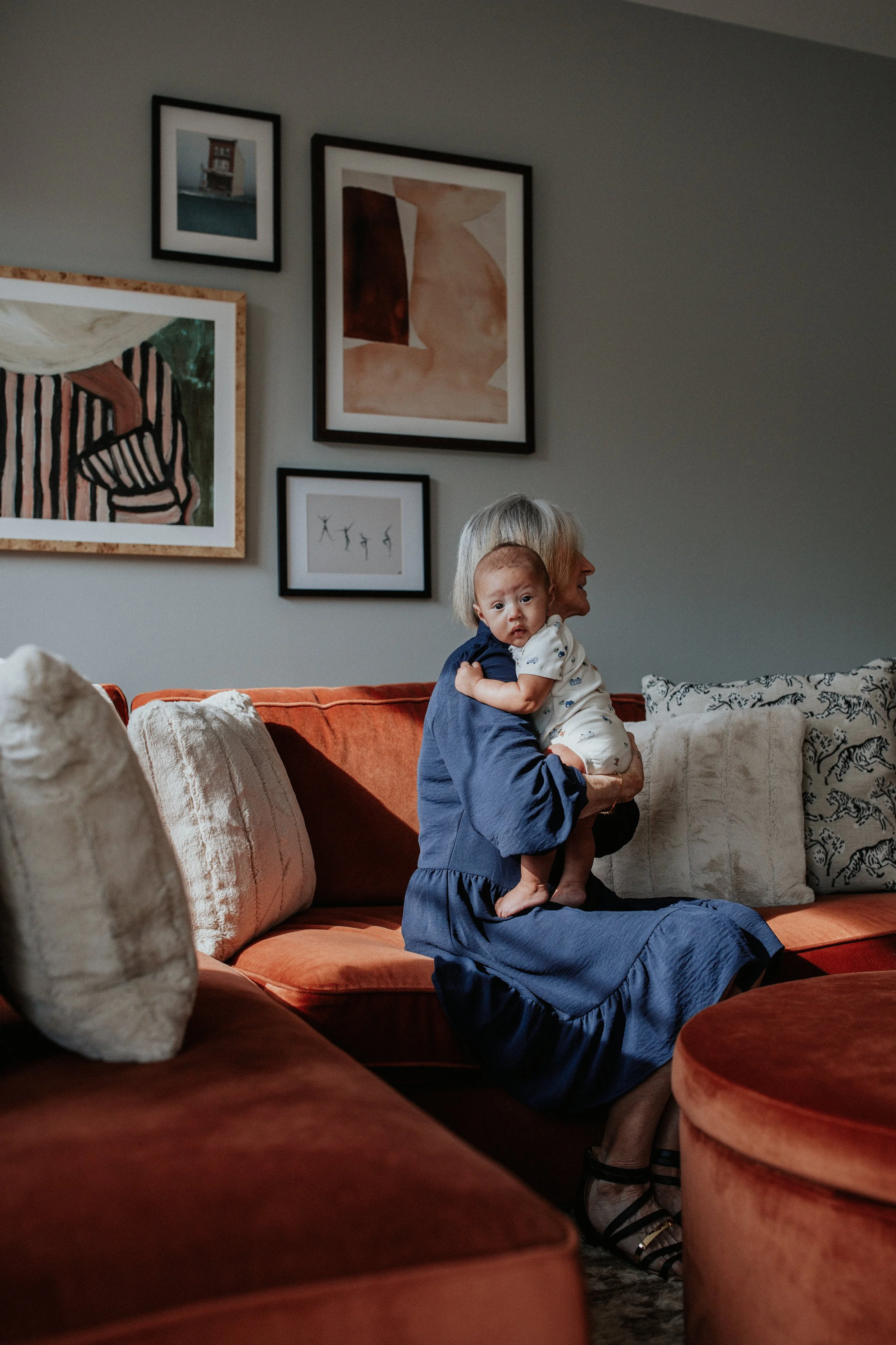 A woman with gray hair sitting on a burnt orange couch holds a baby wearing a white onesie. The woman is looking to her right, while the baby looks directly at the camera, with a slightly puzzled expression. The background features various framed art