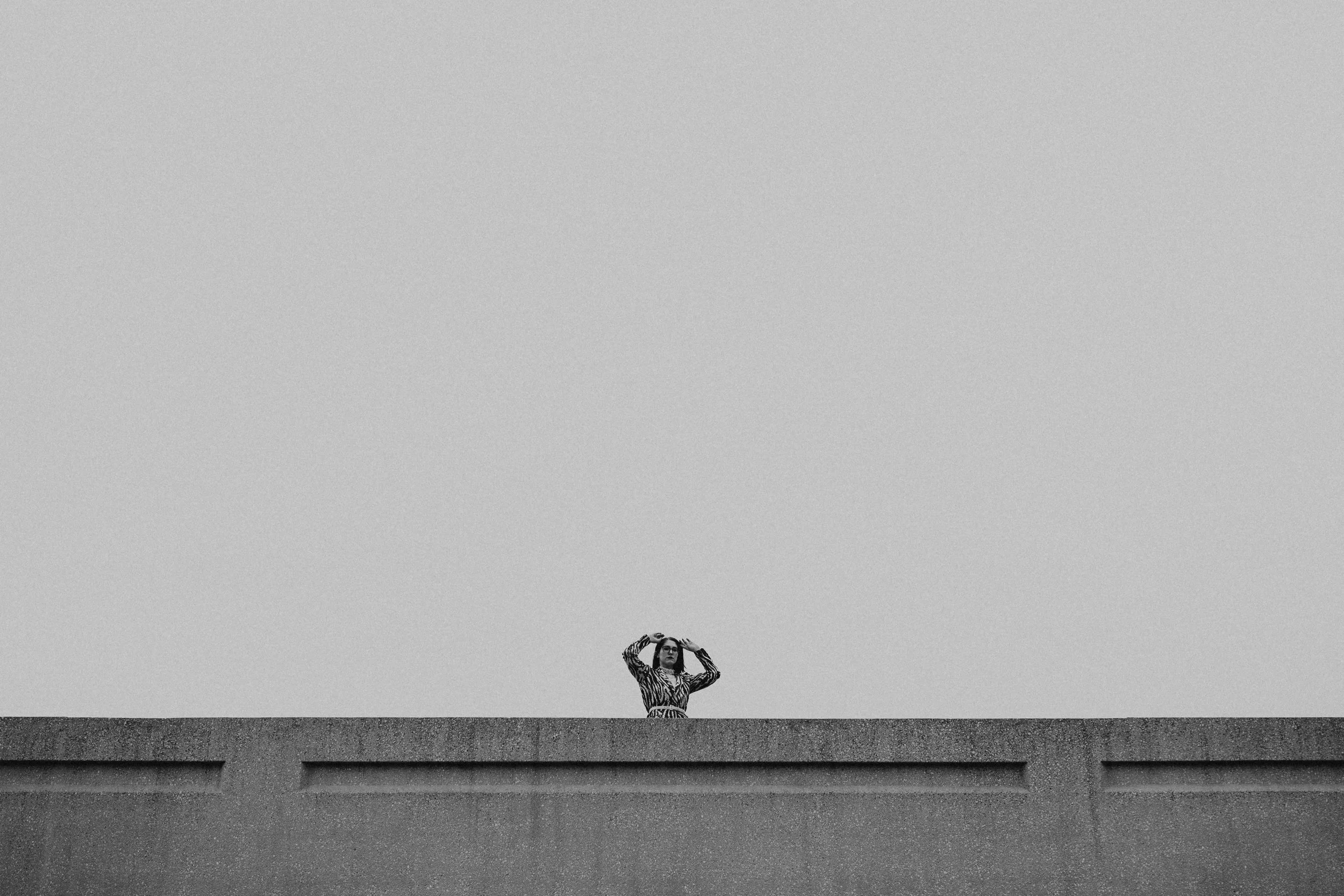 A black and white photo of a woman standing on a ledge, holding her hands above her head, against a stark, clear sky.