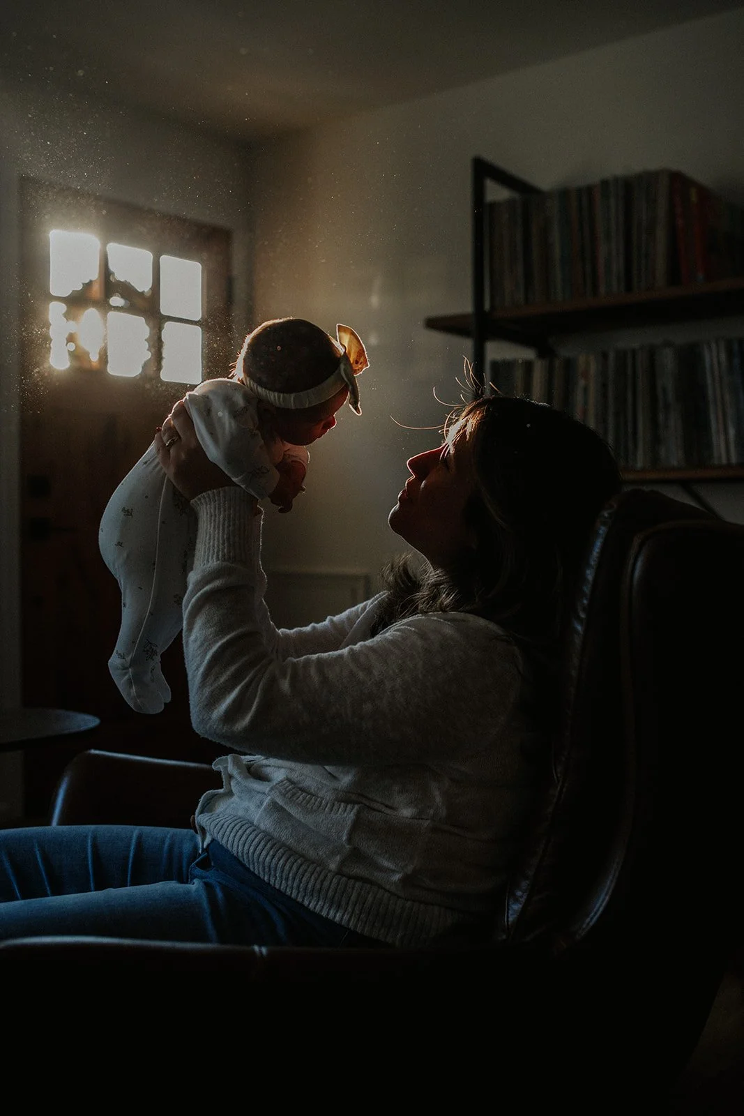 A woman sitting in a dark room, holding a baby up in the air, with sunlight coming through a window behind them.