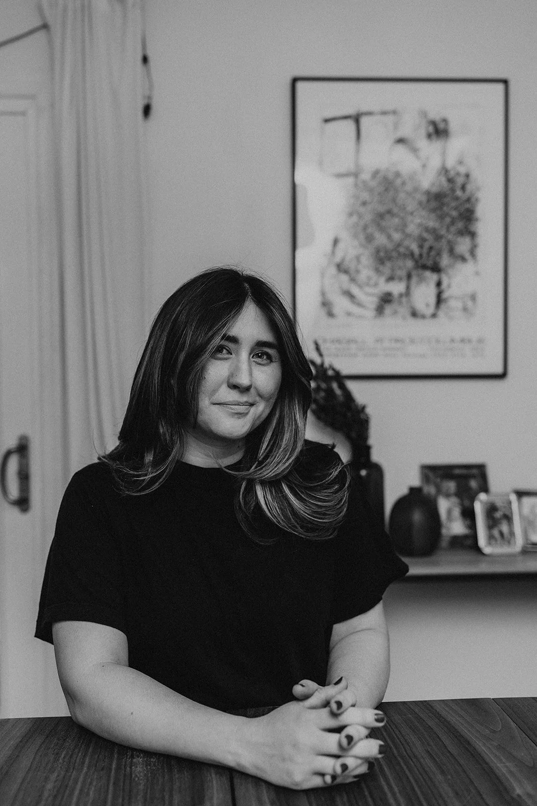 A woman with shoulder-length wavy hair sits at a wooden table, smiling softly. In the background, there is a framed picture of a tree on the wall, a shelf with picture frames, a potted plant, and a vase.
