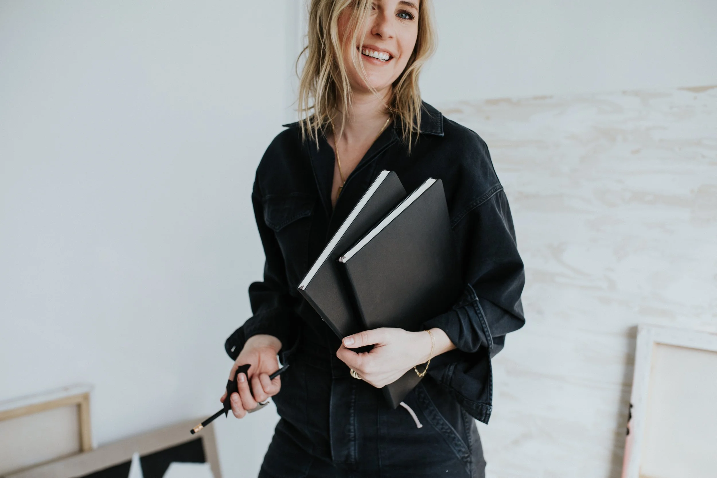 A woman smiling, holding black notebooks and an audio cable, wearing a black jacket, in a room with a white brick wall background.