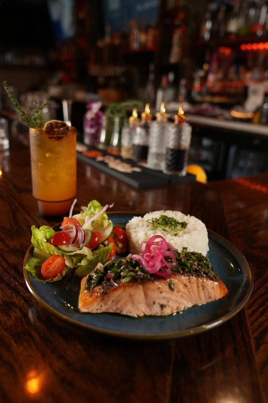 Plate with grilled salmon, rice, and salad on a wooden bar, with cocktail and bar bottles in the background.