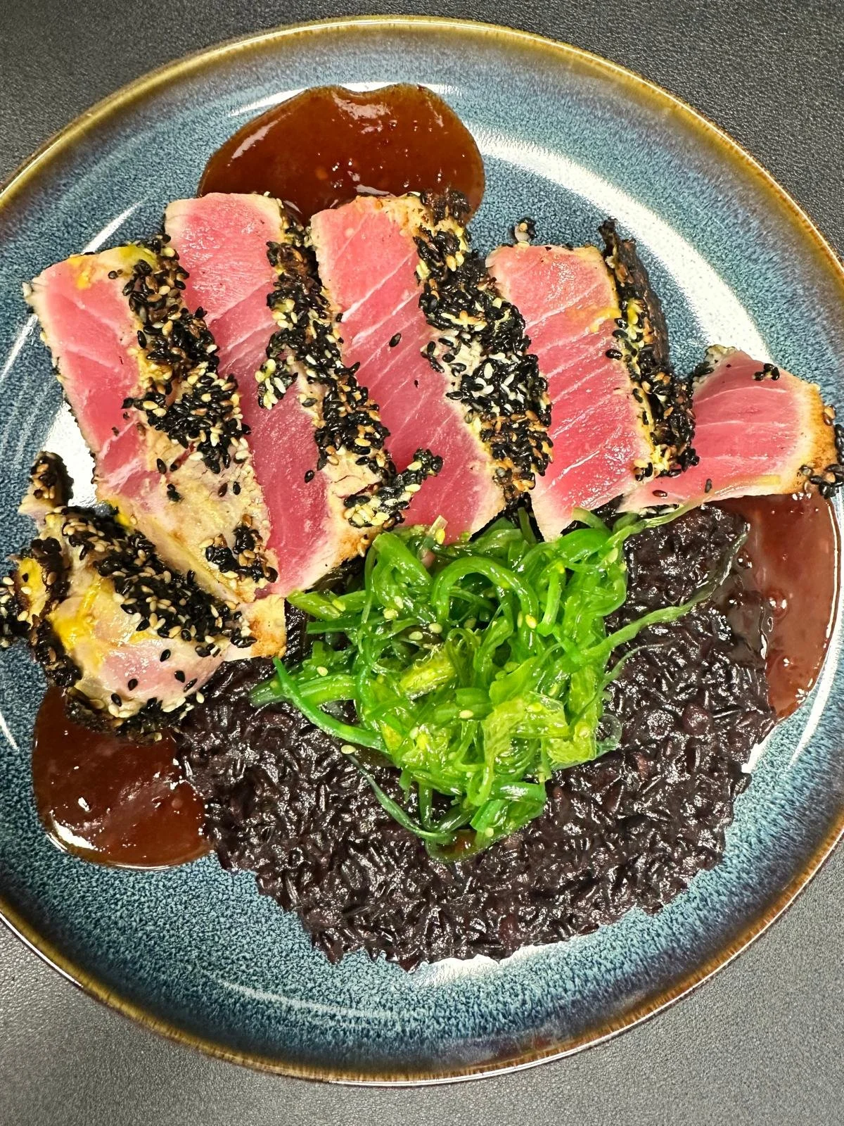Seared tuna coated with black and white sesame seeds, served with green seaweed salad, black rice, and dipping sauce on a blue ceramic plate.