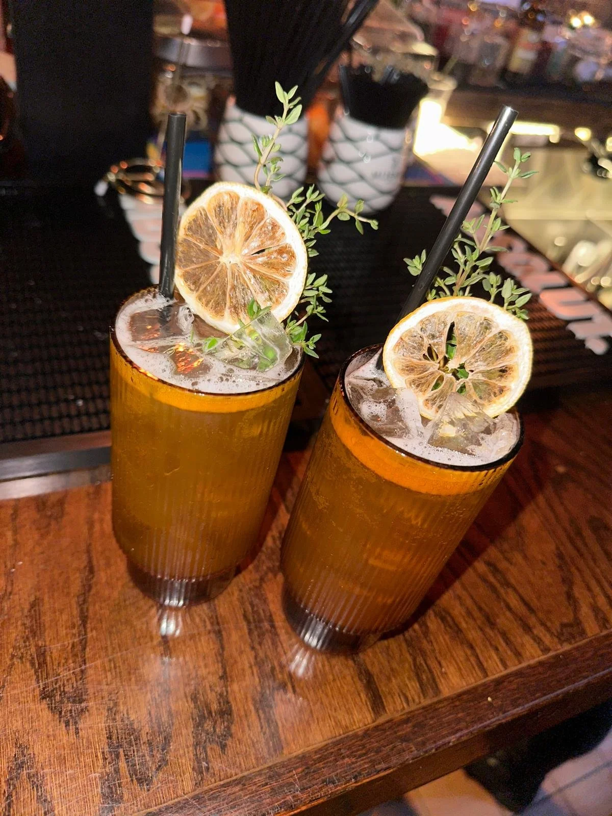 Two tall glasses filled with amber-colored drinks, garnished with lemon slices, sprigs of herbs, ice, and black straws, on a wooden bar counter.